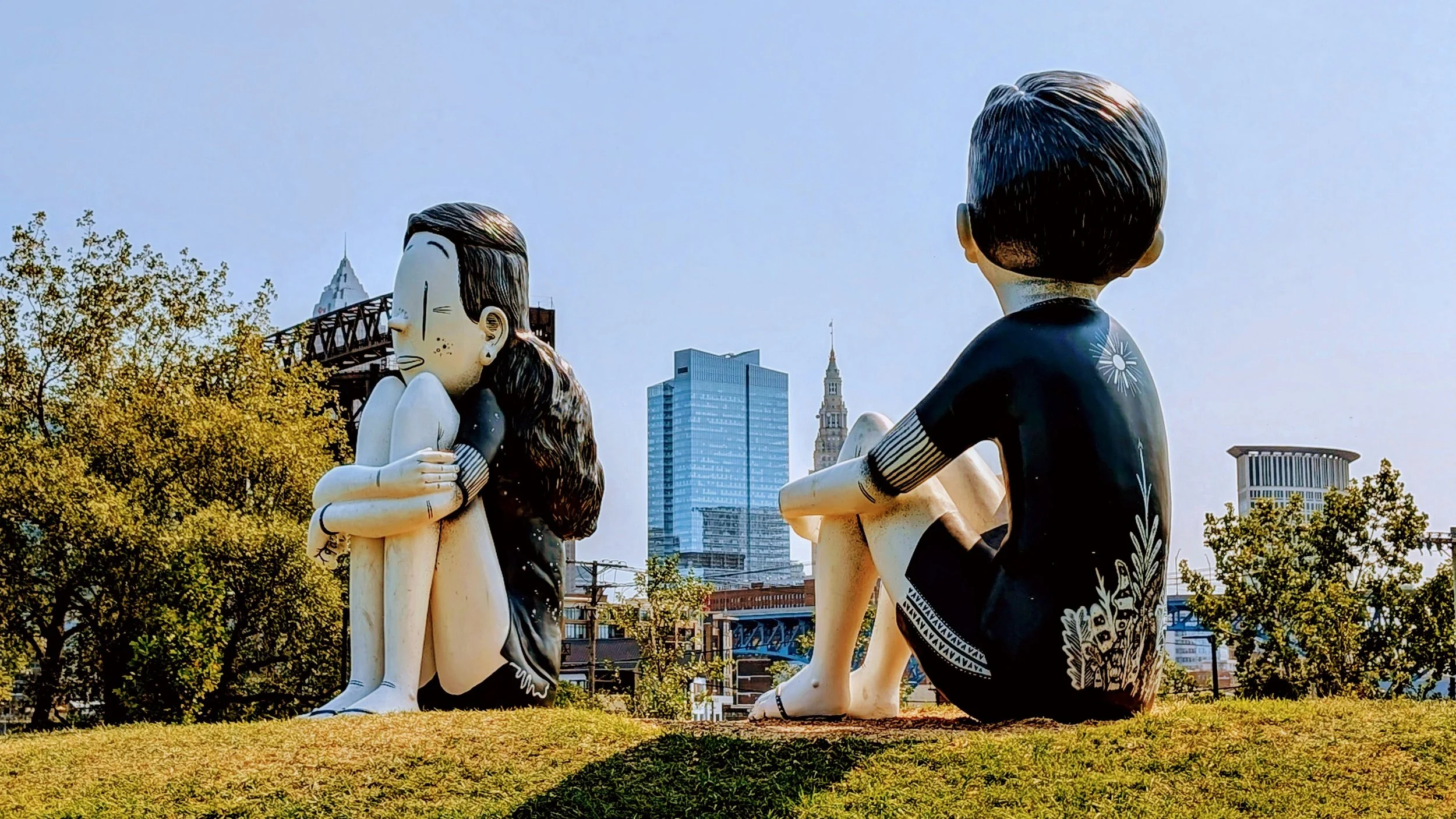 Colorful sculptures of a boy and a girl sitting on grass in front of city buildings, with trees and a blue sky.