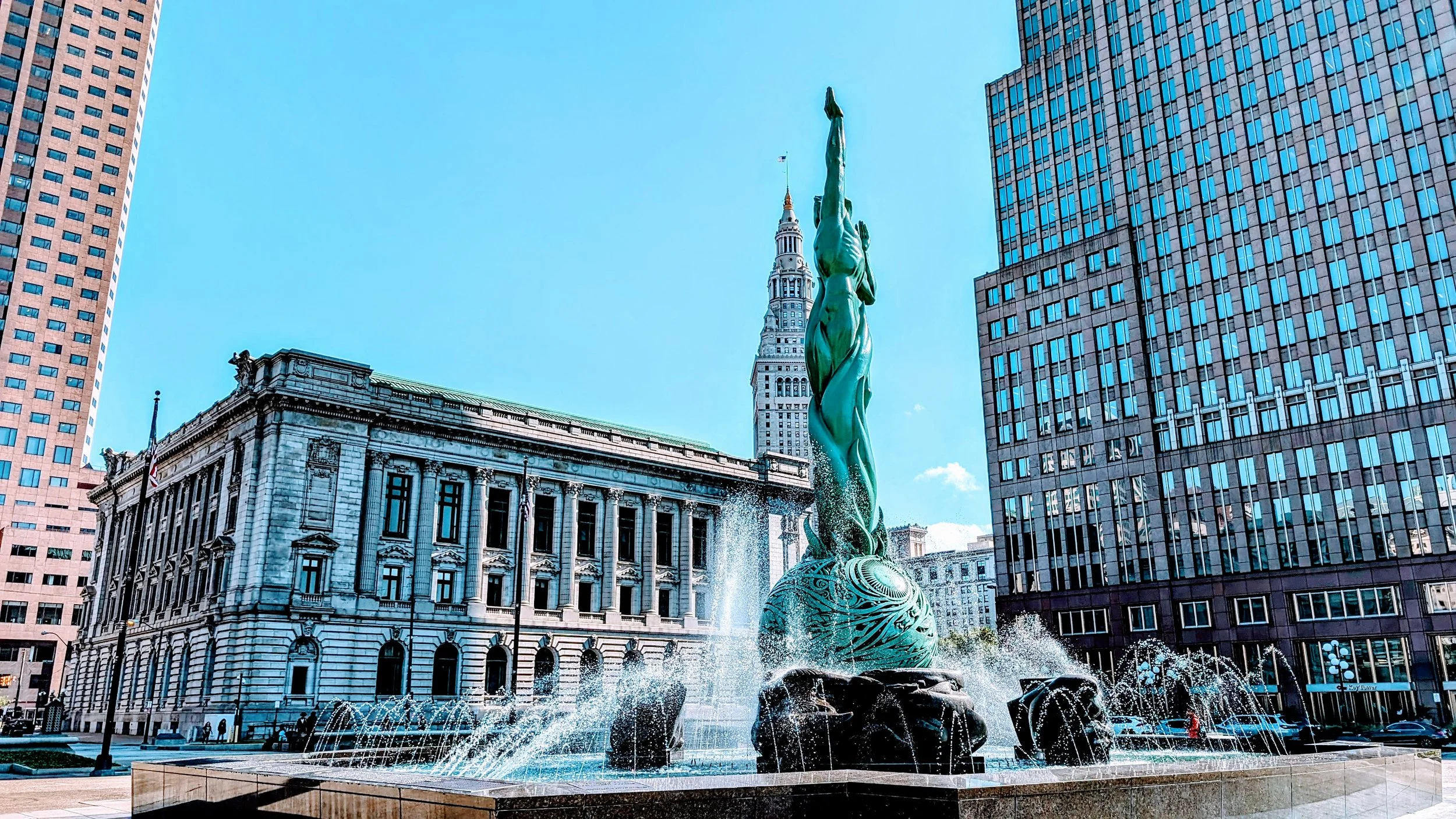 The image features a prominent green fountain sculpture surrounded by water jets, with historic and modern buildings in the background, including the iconic MetLife Tower with its clock face in the distance. The sky is bright and clear.