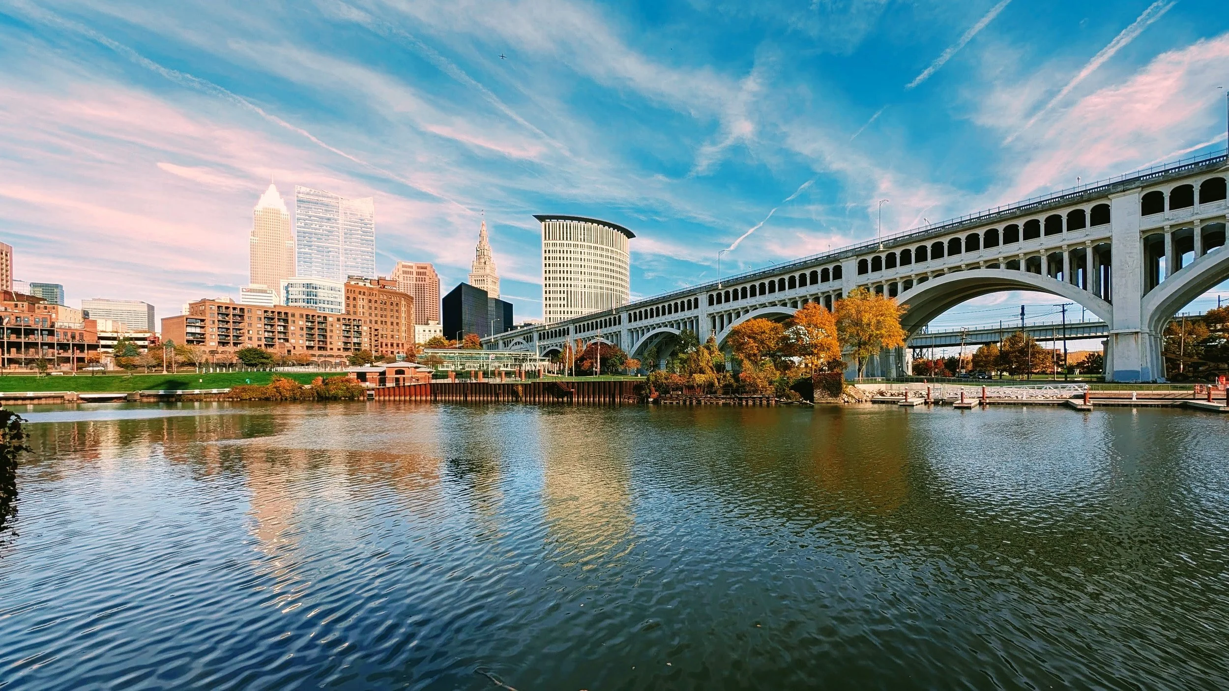 City skyline with high-rise buildings, a large white bridge, and a river in the foreground under a blue sky with wispy clouds.
