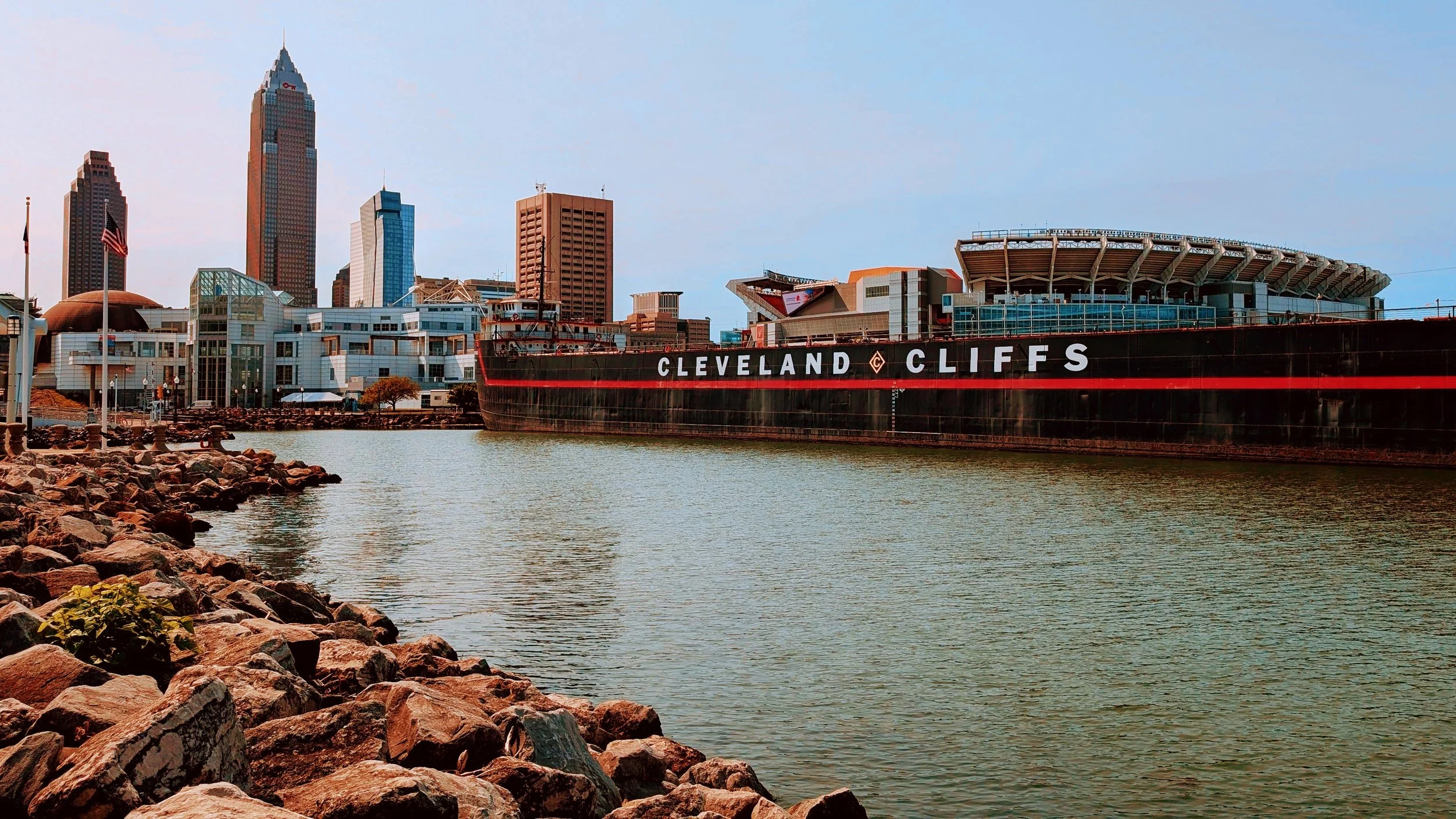 City skyline with tall buildings near a body of water, a large black ship with 'CLEVELAND CLIFS' printed on its side, and a rocky shoreline in the foreground.