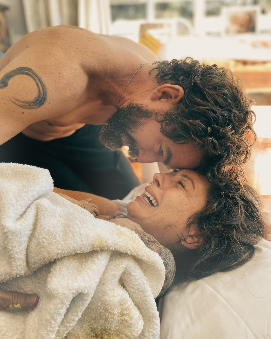 A man with curly hair and a beard leans over a woman lying on a bed, smiling and showing affection. The woman holds a new born baby. Both woman and baby are wrapped in a white blanket.