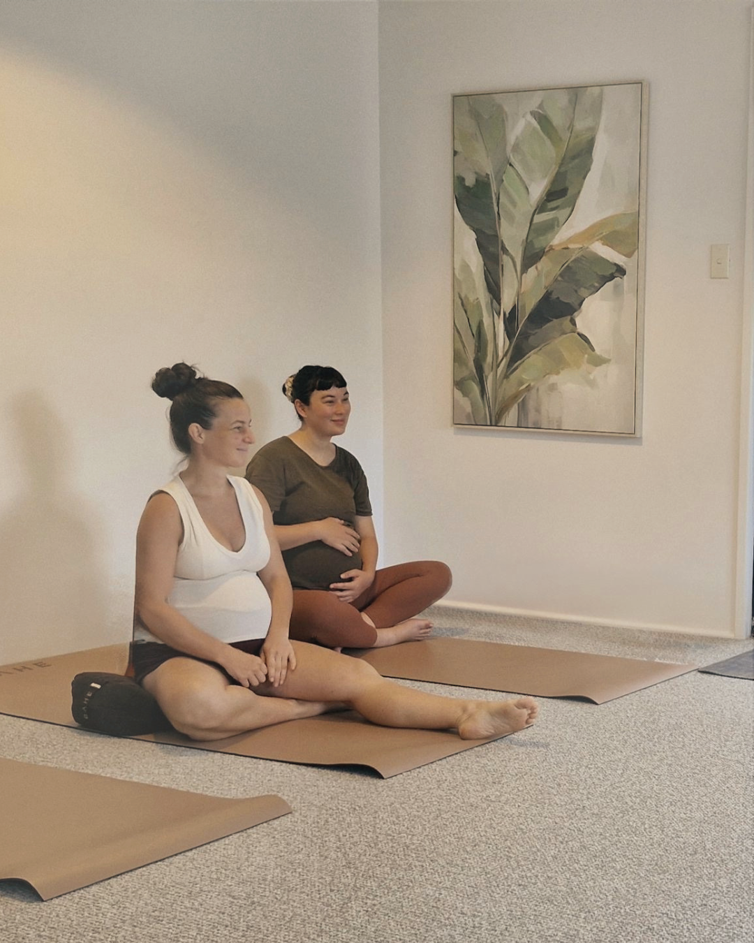 Two women practicing yoga in a room with a large leafy painting on the wall, sitting on yoga mats.