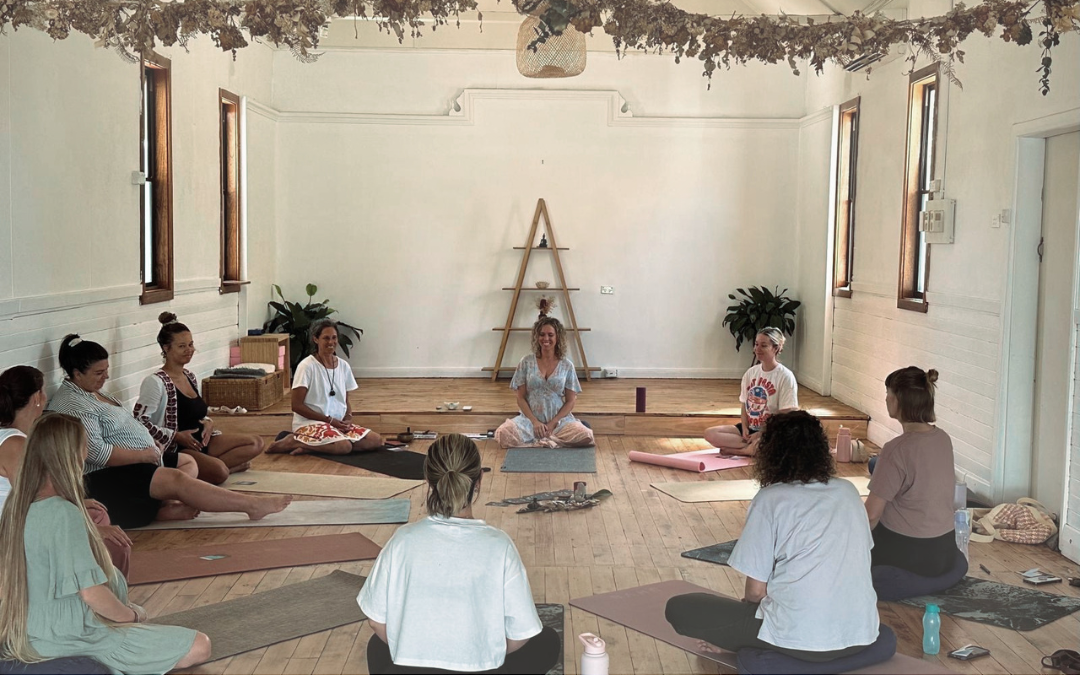 Group of women practicing yoga or meditation in a bright, airy room with wooden floors, natural light from windows, and decorative elements.