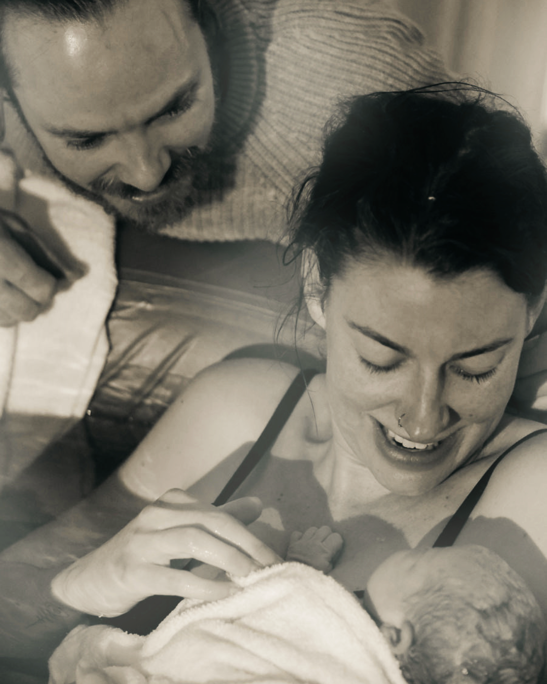 A woman in a birthing pool, holding her newborn baby wrapped in a towel, smiling with eyes closed, and a man leaning over her, smiling and looking at the baby.
