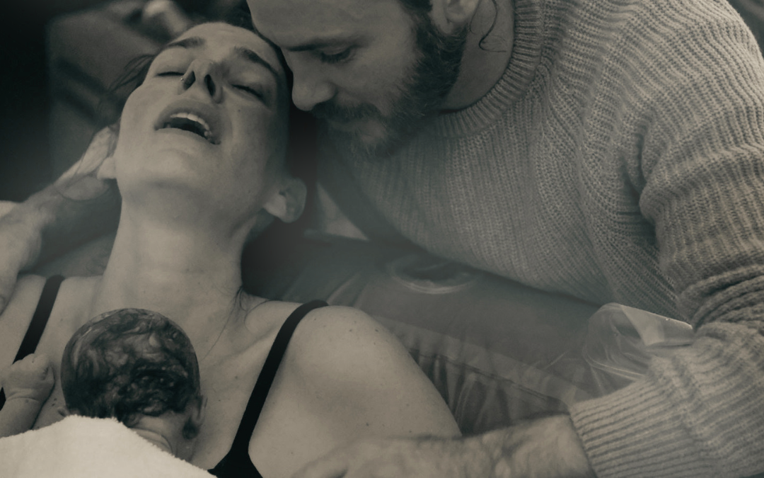 A woman holding a newborn baby on her chest after birth, her husband leaning over her, arms wrapped around his wife and baby, their heads touching gently.