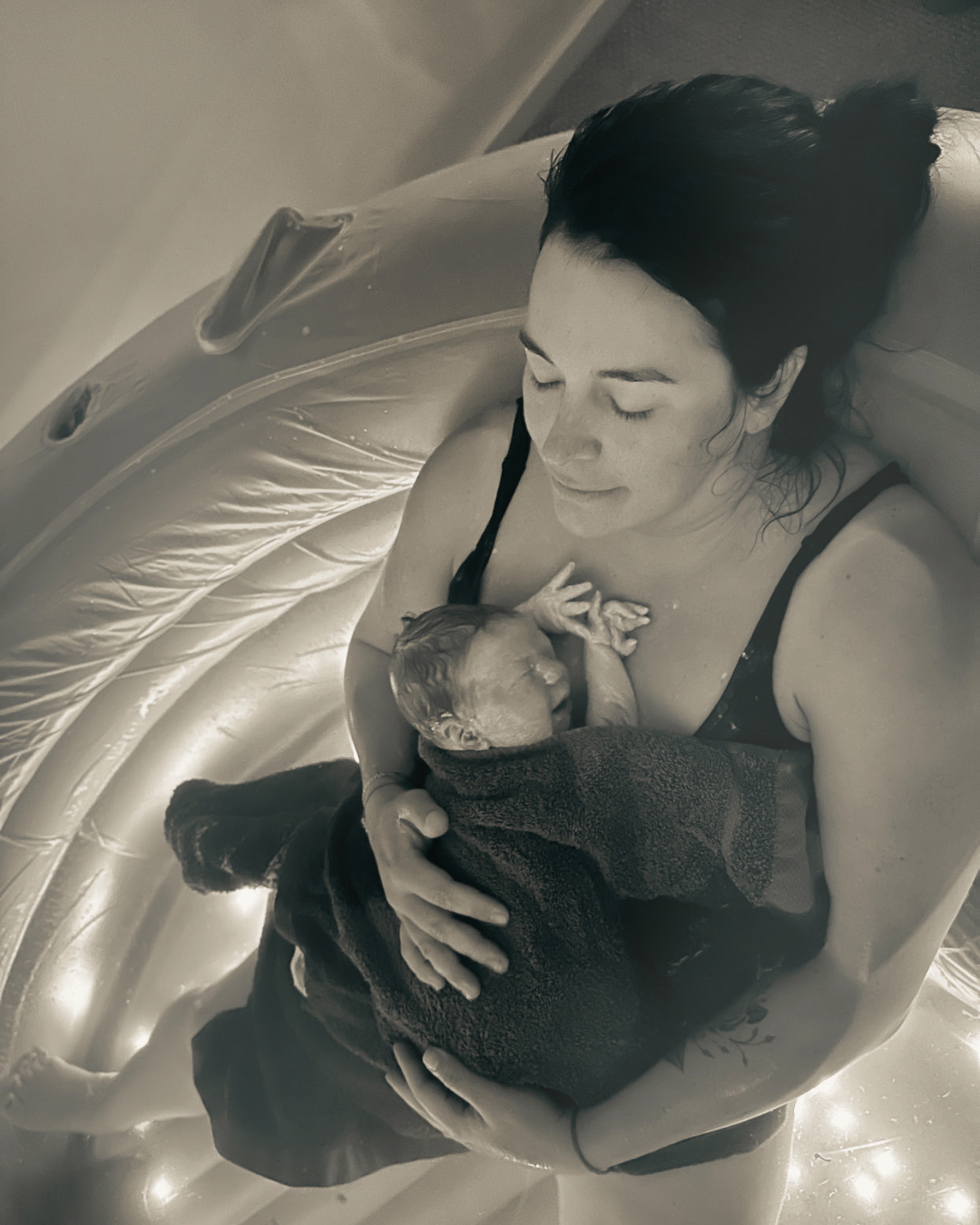 A woman lying in a birthing pool, holding a newborn baby wrapped in a dark towel, both appearing peaceful and content.
