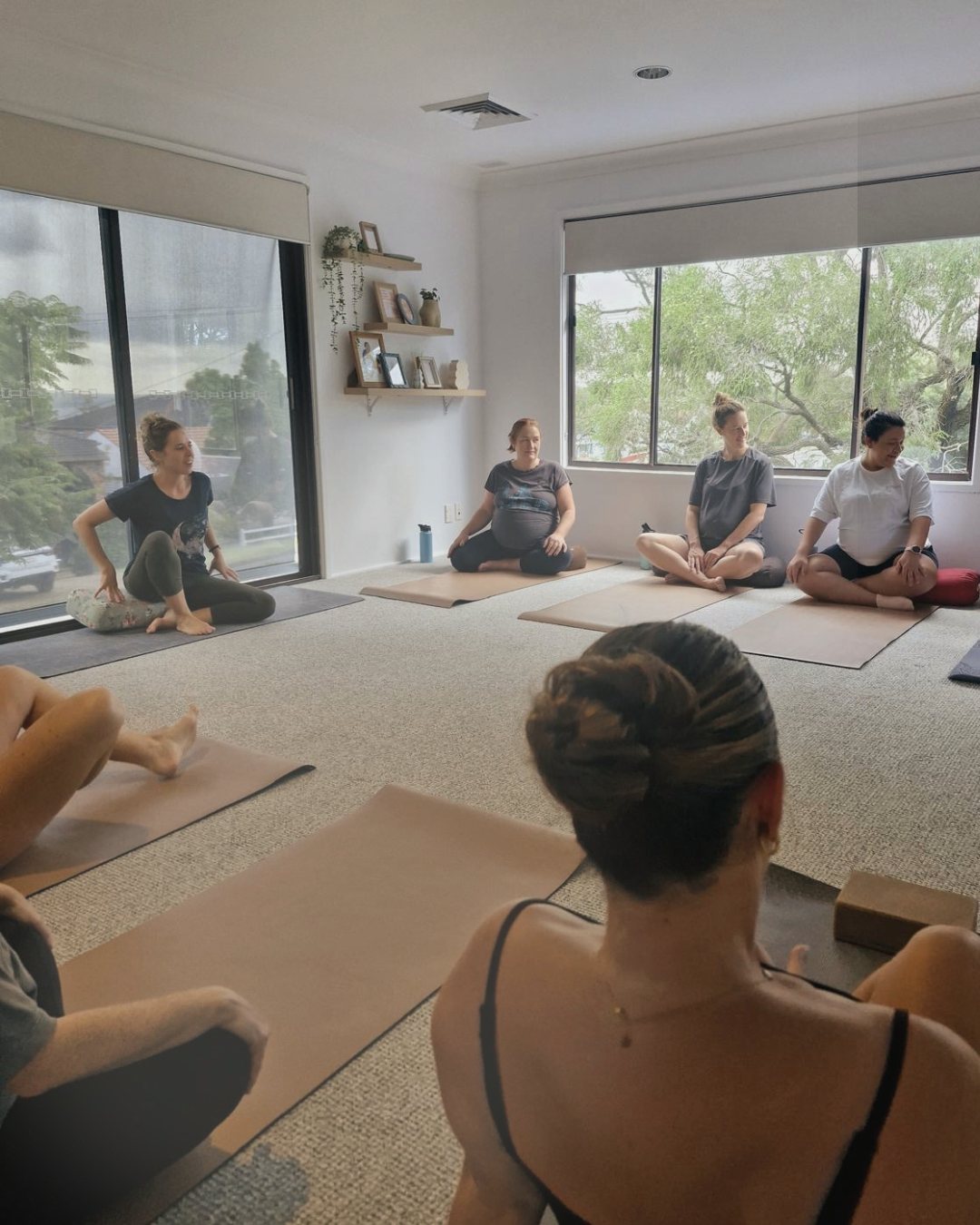 Group of women participating in a yoga class in a bright, airy room with large windows and minimalist decor.