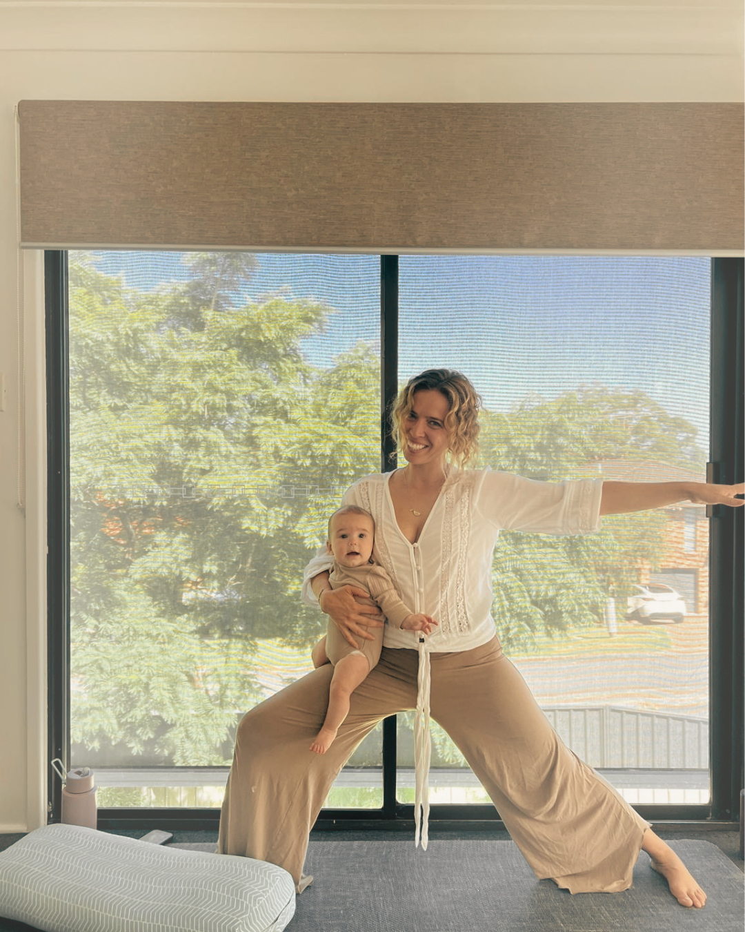 A woman with curly blonde hair smiling and practicing yoga in front of a large window, holding a baby in her arms inside a room with beige curtains and a small pillow on the floor.