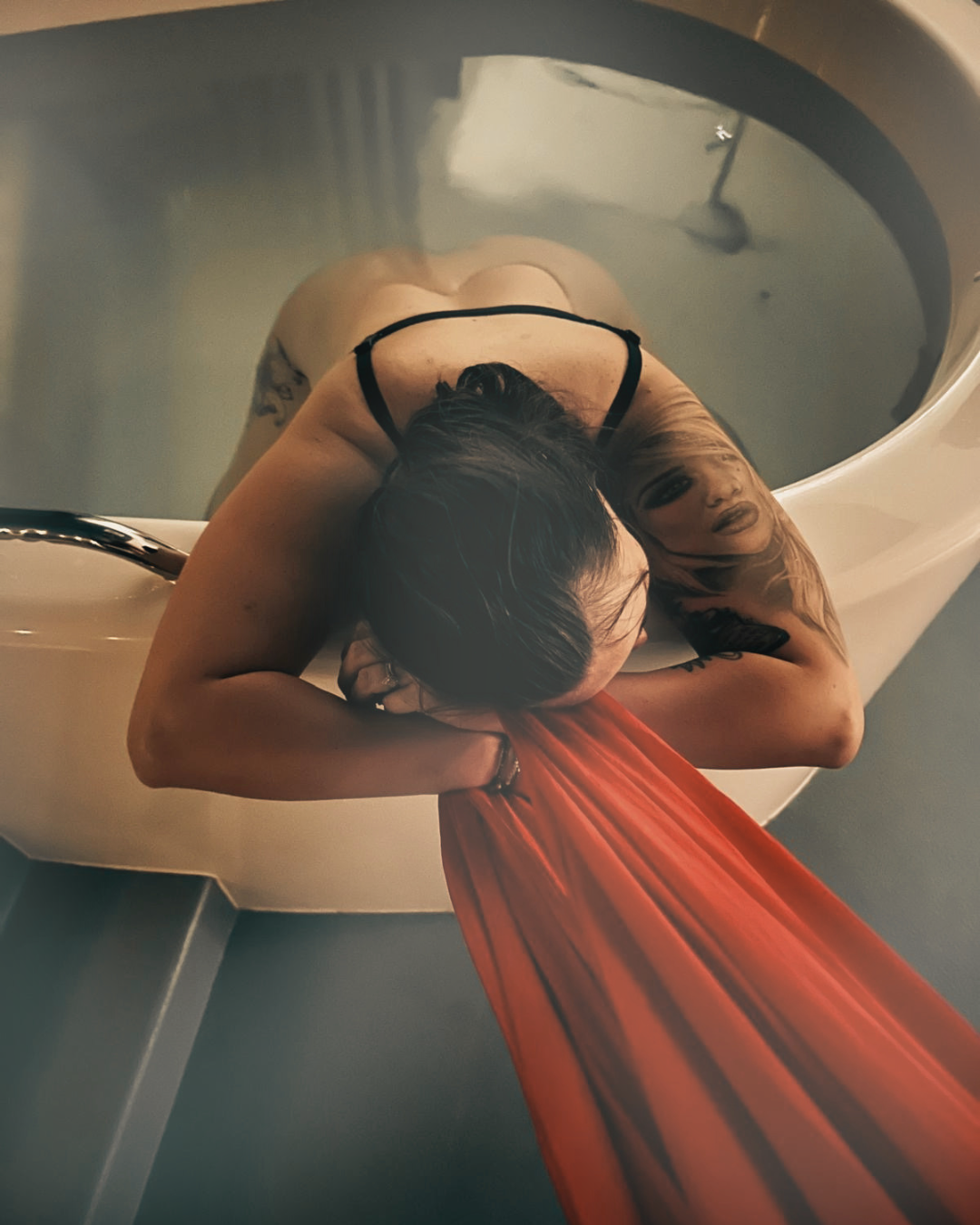 A woman with dark hair is lying on the edge of a birthing pool, arms folded, head down and holding some red fabric.