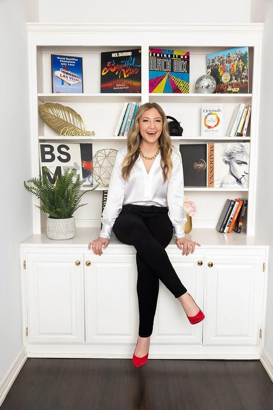 A woman sitting on a white cabinet in front of a white bookshelf, smiling, wearing a white blouse, black pants, and red high heels, with books, decor, and a potted plant on the shelves.
