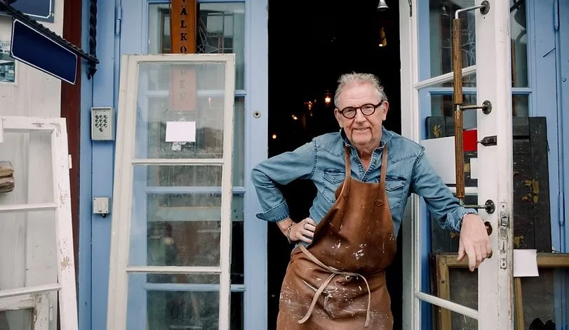 An elderly man with gray hair and glasses stands at the entrance of a store or workshop, wearing a denim shirt and a leather apron with paint or stain marks, smiling and leaning on the doorframe.