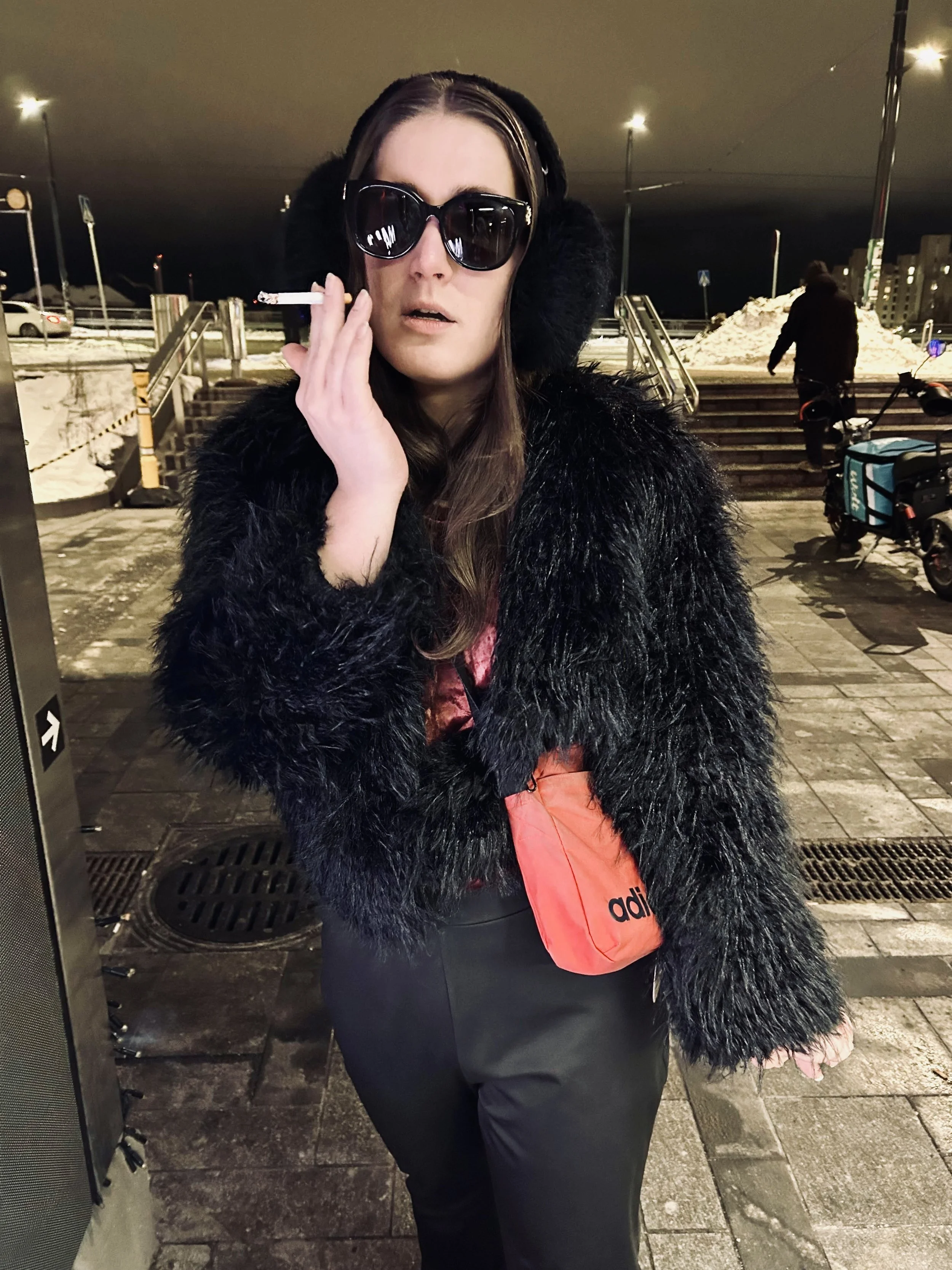 A woman in dark sunglasses and a black faux fur coat standing outdoors at night, holding a cigarette in her right hand near her face, with snow and steps in the background.