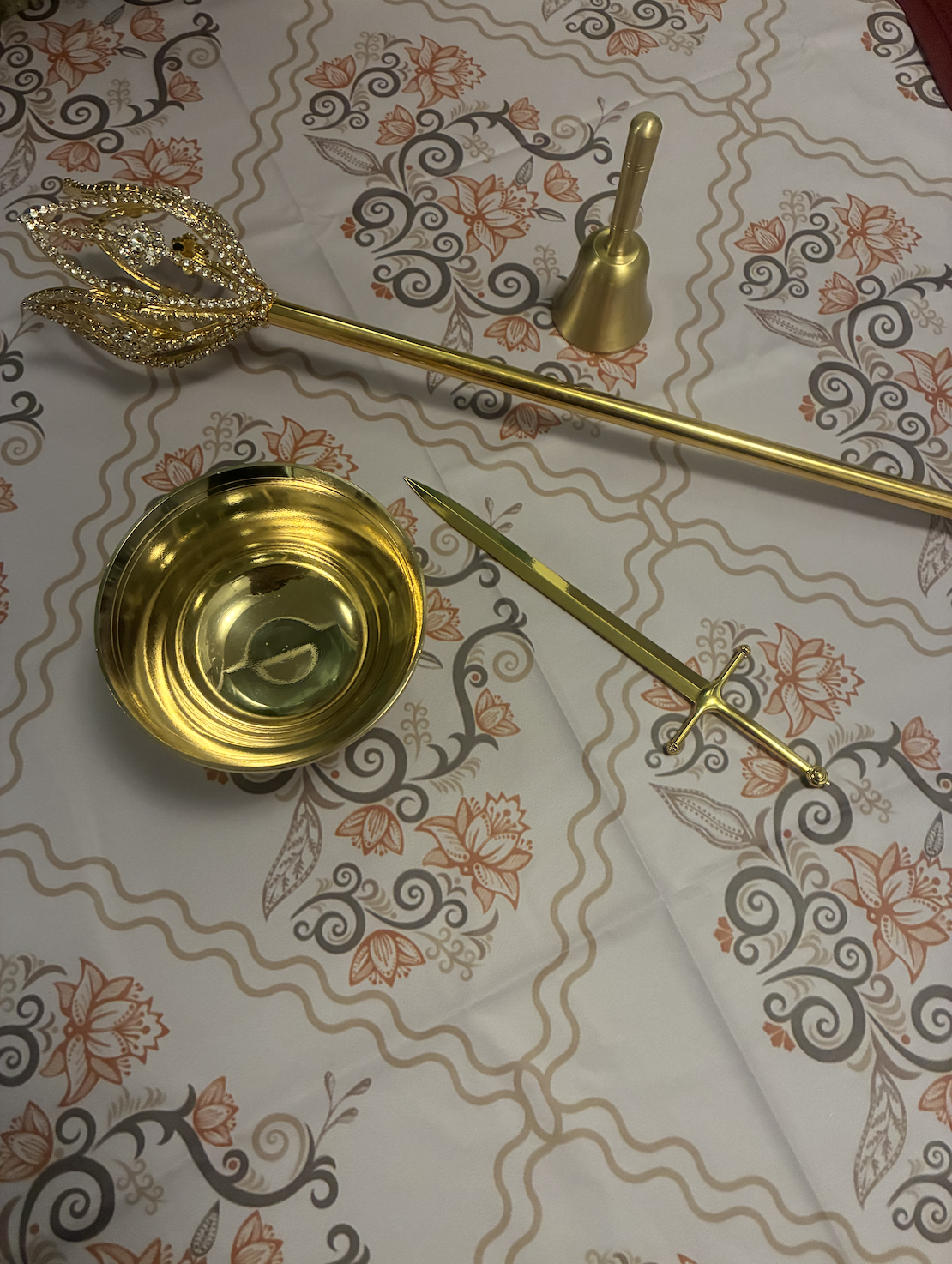 A gold-colored ceremonial set on a tablecloth with a floral pattern. The set includes a wand, a small cup, a dagger, and a bell.