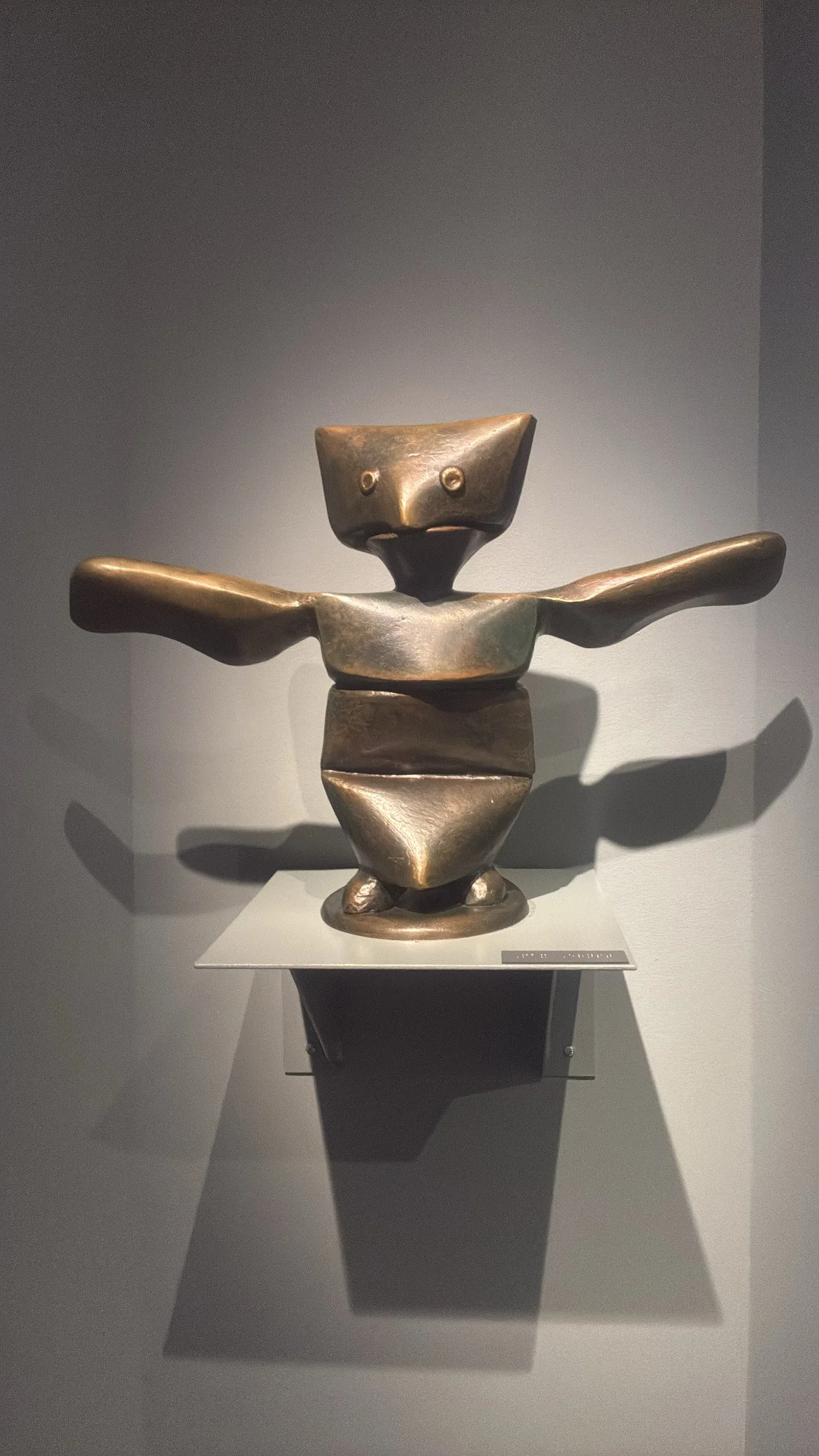 Bronze abstract sculpture of a cartoonish figure with outstretched arms, a square head with two small round eyes, and a rounded body, displayed on a white pedestal against a grey wall.