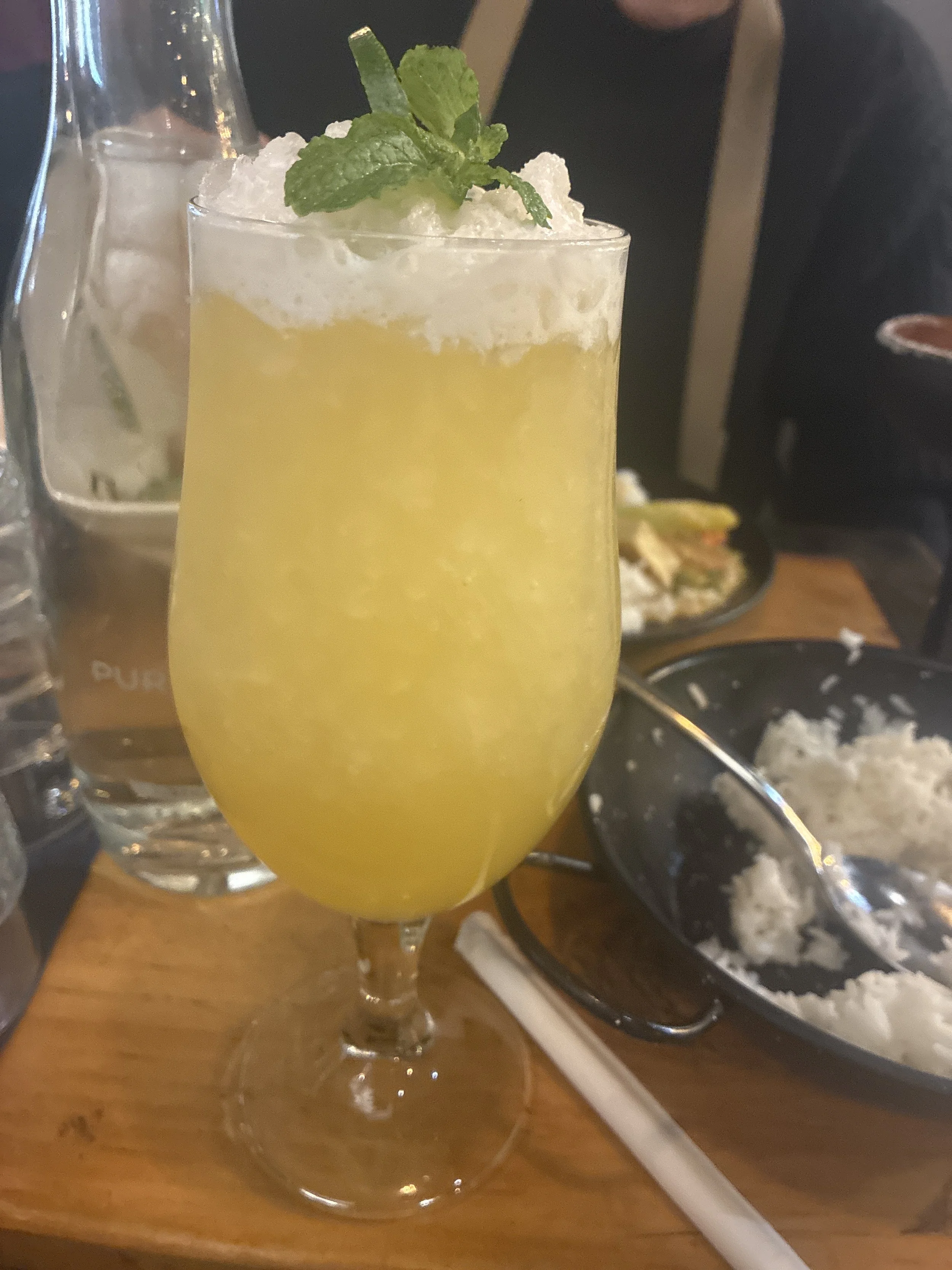 A tall glass of yellow drink garnished with mint leaves, topped with whipped cream or foam, on a dining table with rice and other dishes in the background.
