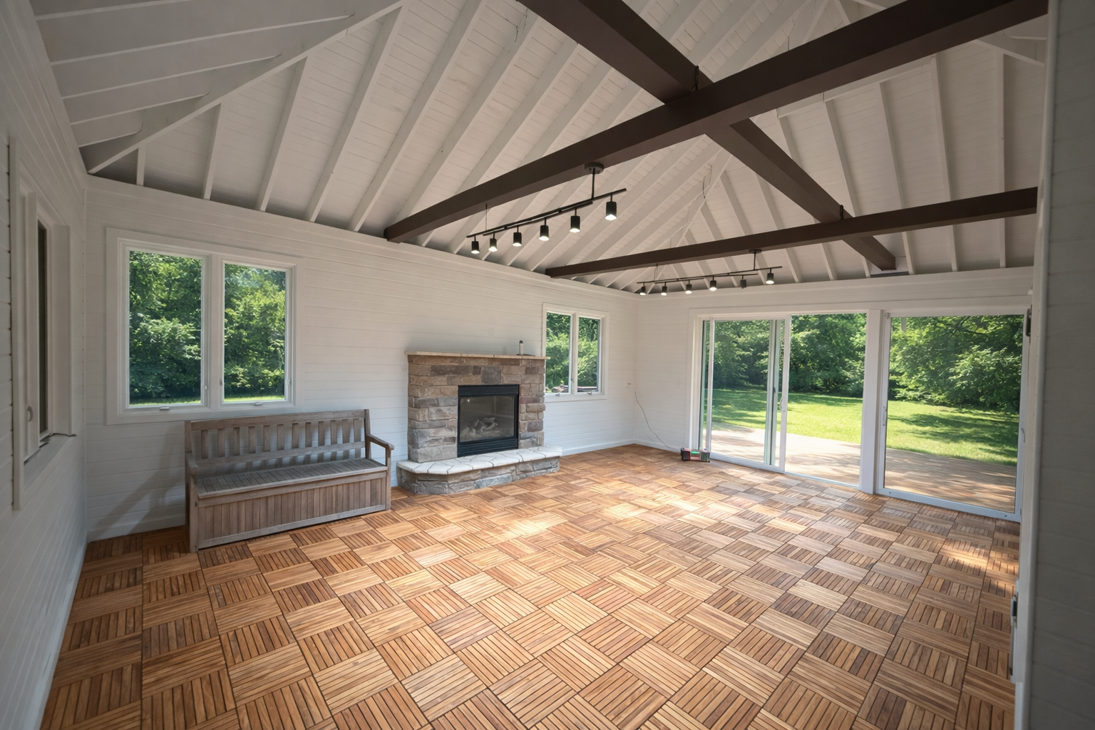 Empty sunroom with vaulted white ceiling and exposed dark beams, large windows, tiled hardwood floor, stone fireplace, wooden bench near window, sliding glass door leading outside to green lawn.