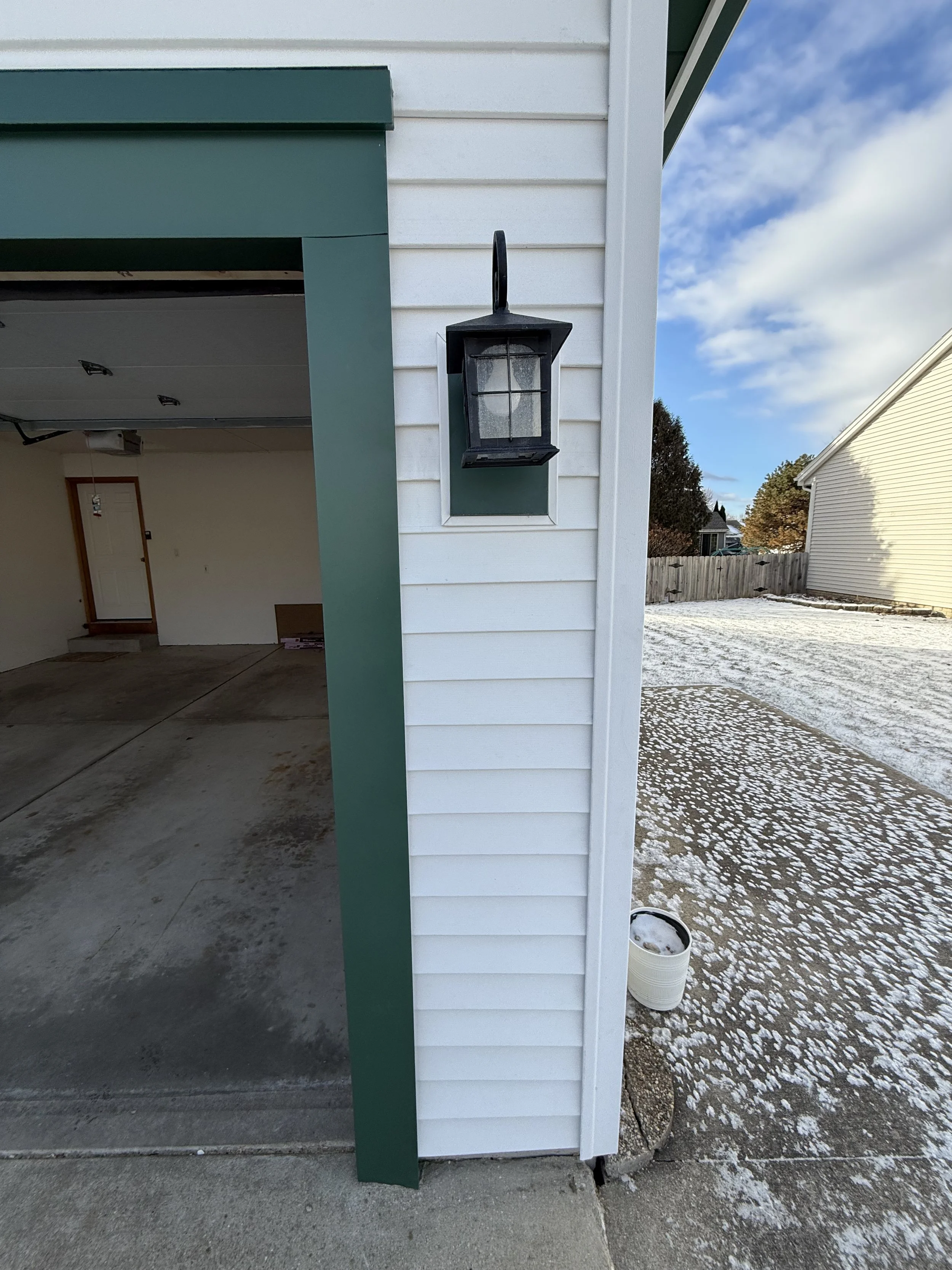 Side view of a garage with white siding, green trim, and a black outdoor lantern attached to the wall, with a partially open garage door revealing an empty interior. Snow is on the ground.