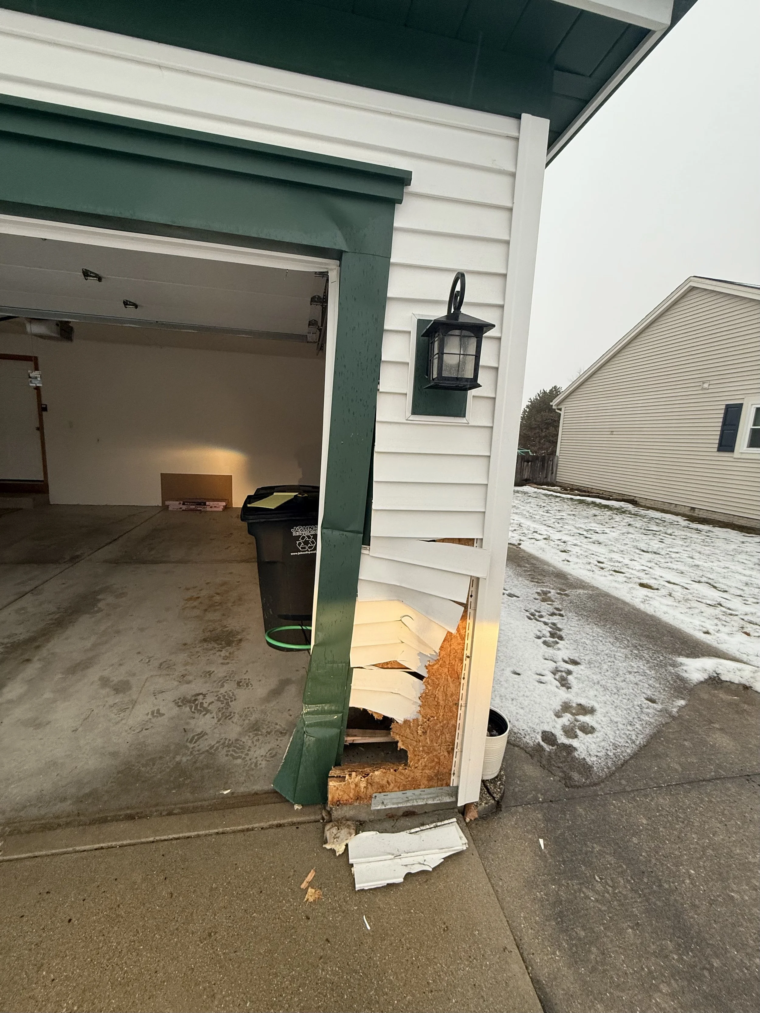 Side of a garage with damage to the siding, exposing wooden structure underneath, and snow on the ground outside.