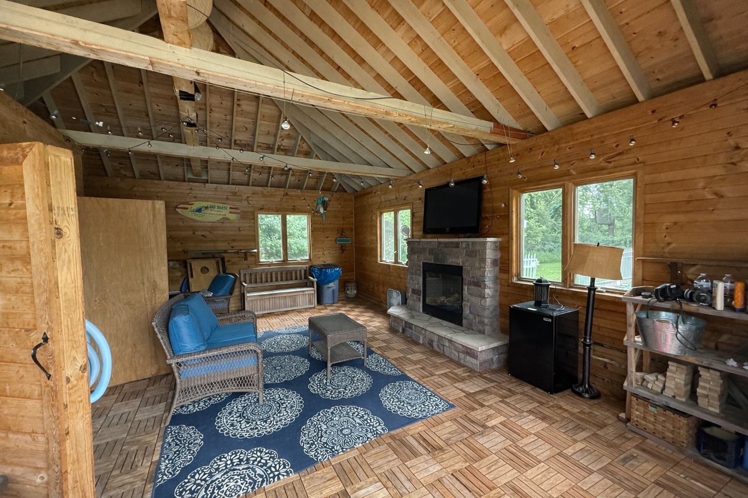 A rustic living room with wooden walls and a vaulted ceiling featuring exposed wooden beams. The room has three windows letting in natural light, a stone fireplace with a flat-screen TV mounted above, a blue patterned rug, wicker and wooden chairs, a small table, and various decorations and furniture.