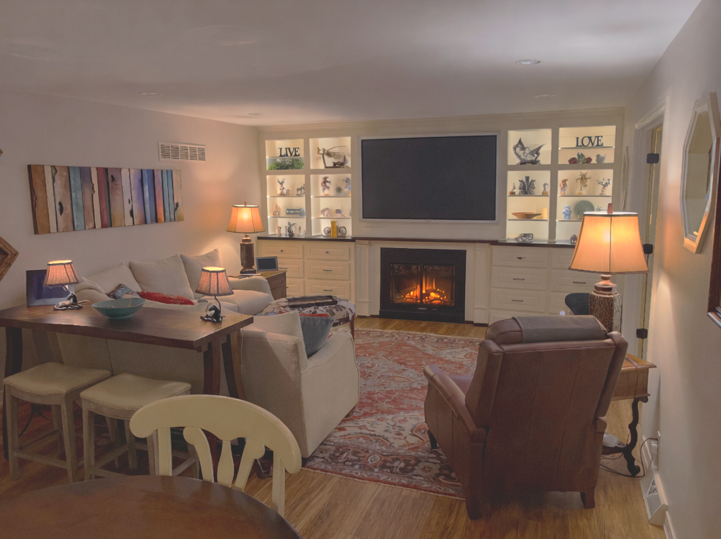 Cozy living room with a fireplace, wall-mounted TV, built-in shelves with decorative items, beige sofa, leather armchair, multiple table lamps, and colorful wall art.