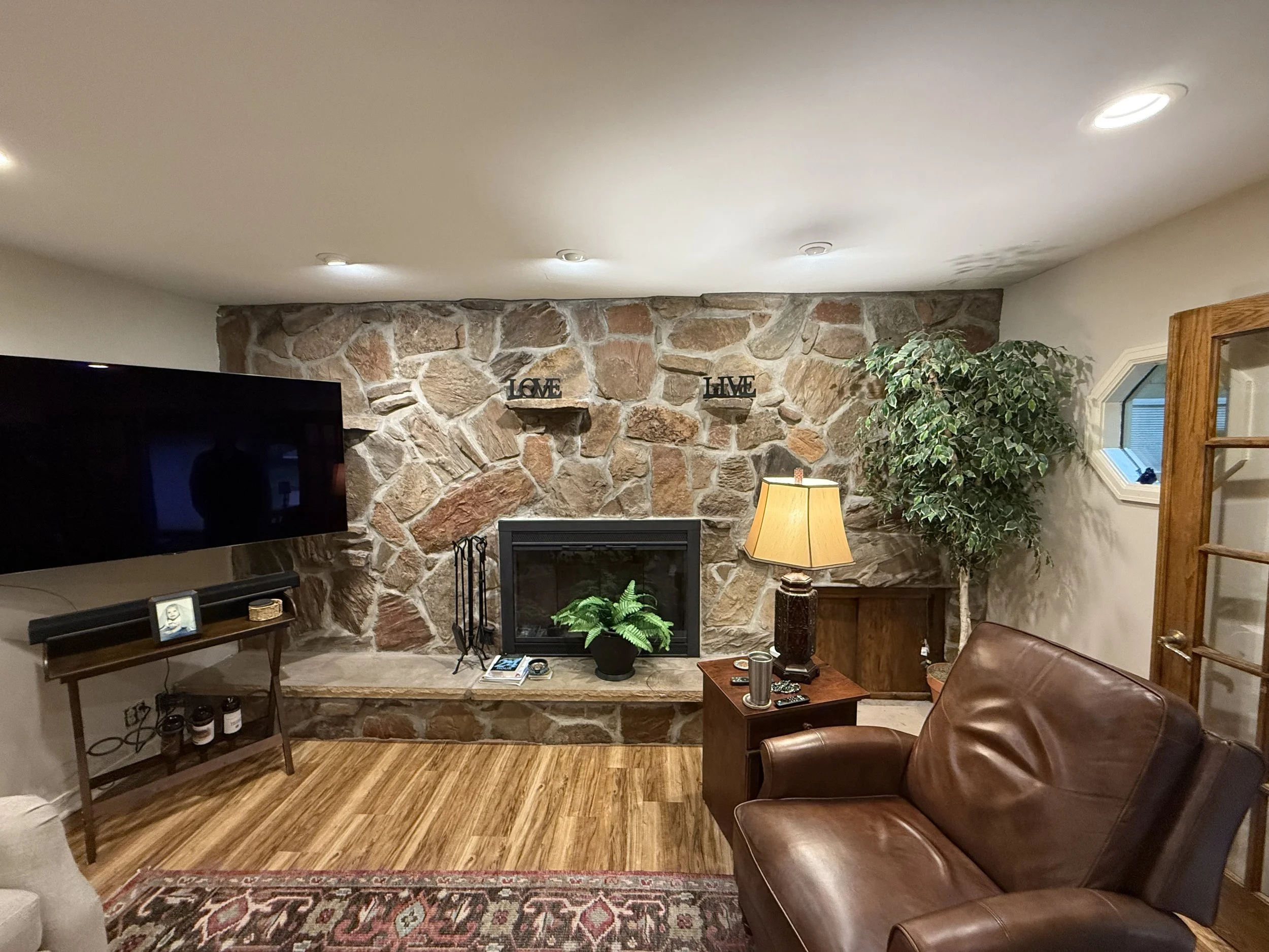 Living room with stone fireplace, large TV to the left, a brown leather couch on the right, a side table with a lamp, a decorative plant, and wall decor that says "LOVE" and "LIVE".