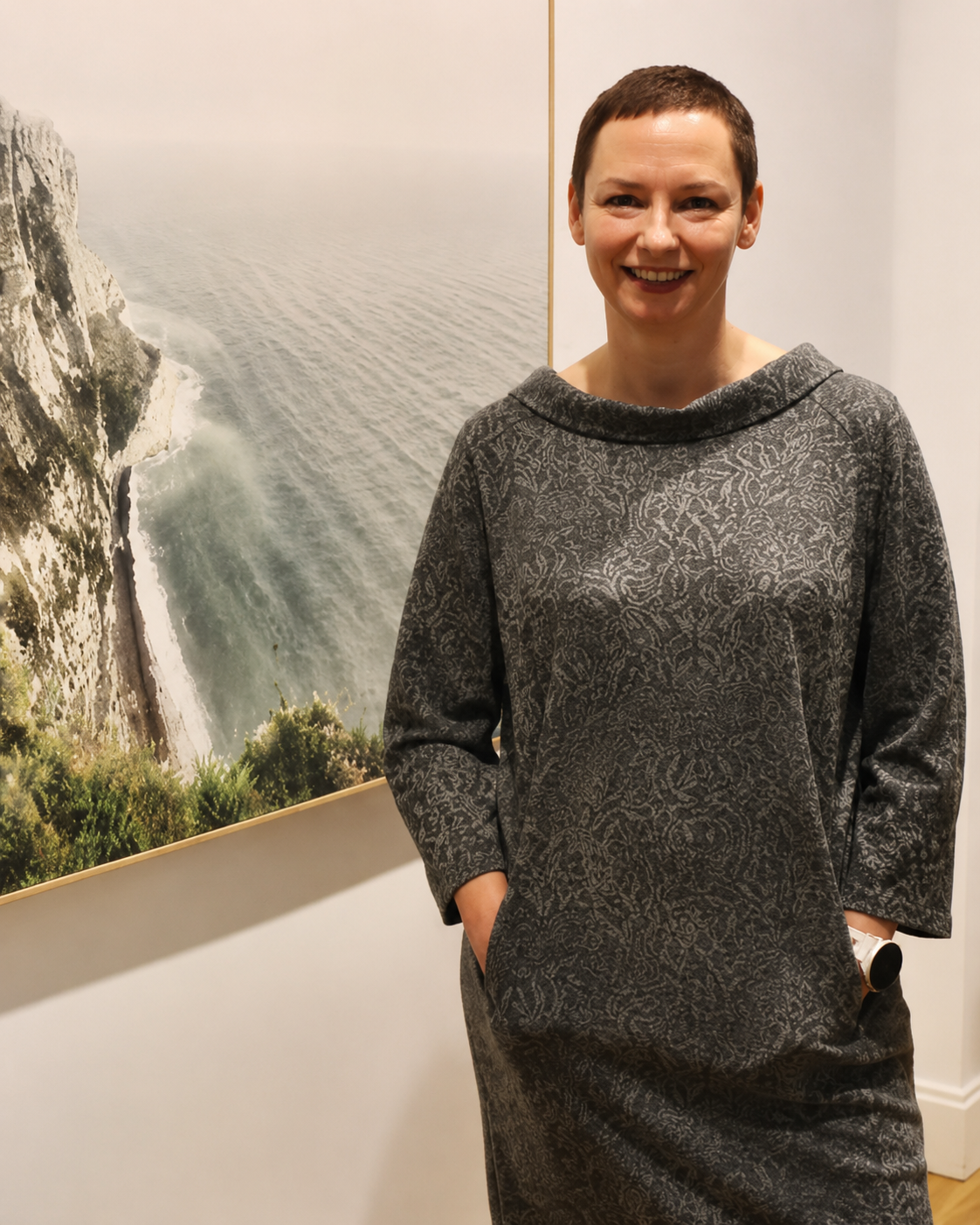 Smiling woman with short brown hair wearing a gray patterned dress standing in front of a large framed photograph of a cliff by the ocean.