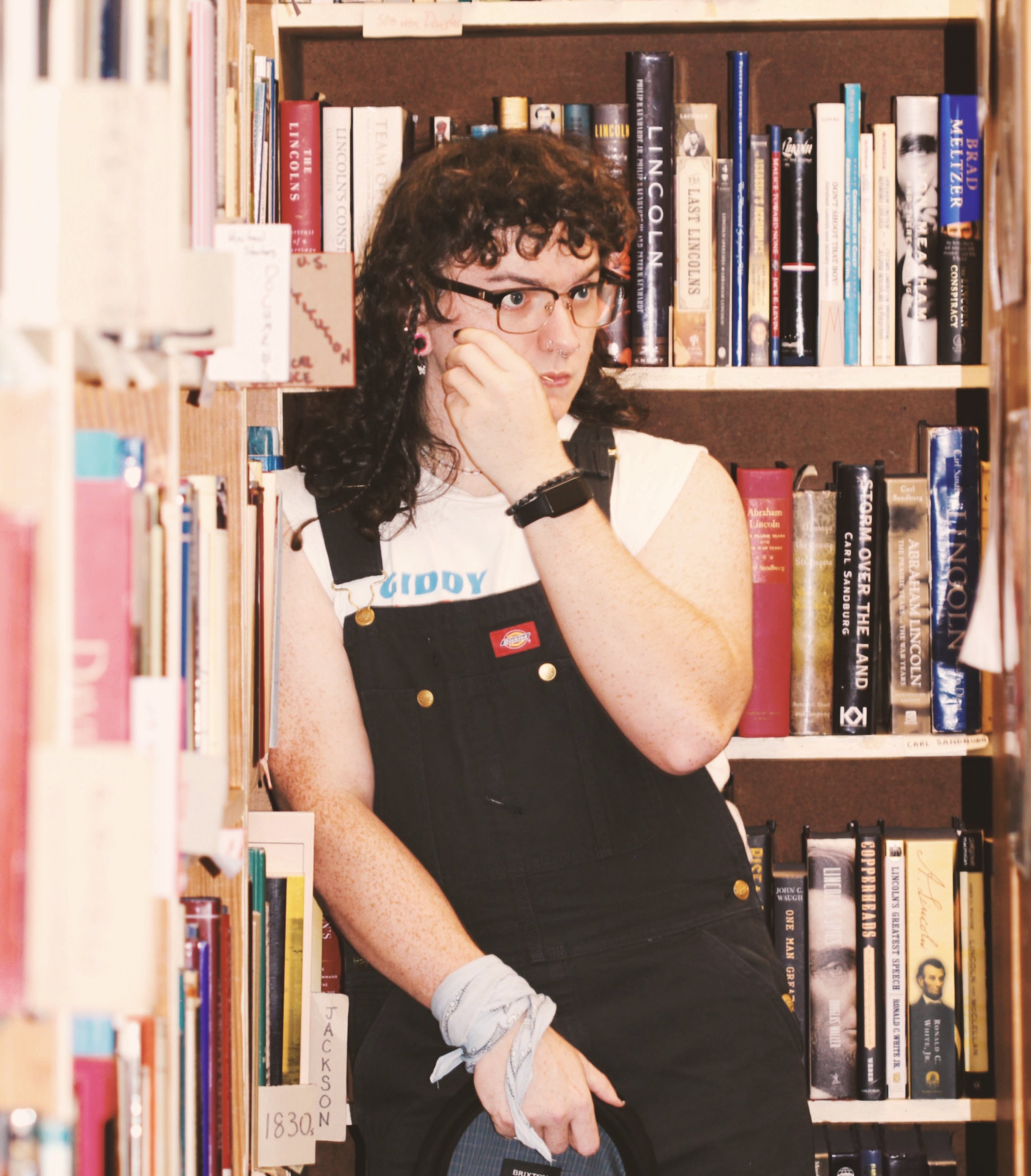 Photo of a woman wearing overalls standing in a library. She has a blue bandana on her wrist, a toothpick in her mouth, long curly hair, glasses, and a toothpick in her mouth. She is looking away from the camera.