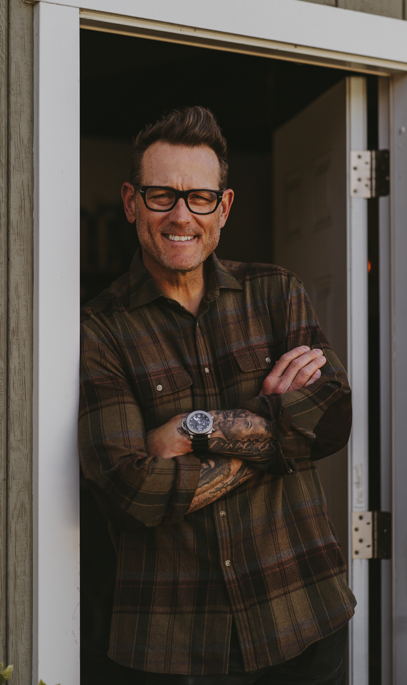 A smiling man with glasses, tattoos on his arm, and a plaid shirt standing in a doorway with arms crossed.