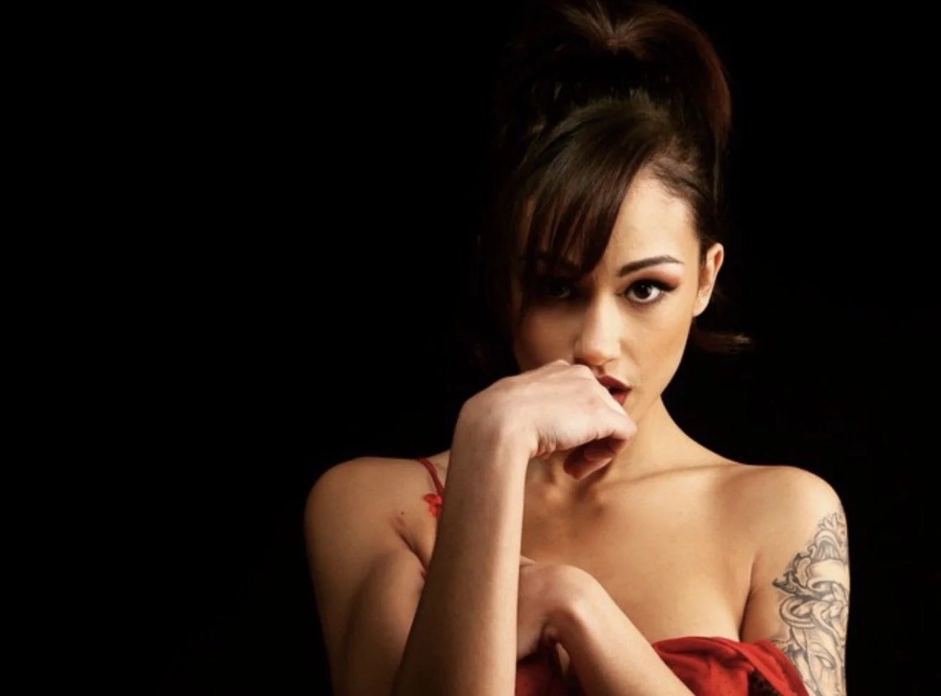 A young woman with dark hair styled in an updo, wearing a red strapless top, covering her mouth with her hand, against a black background.