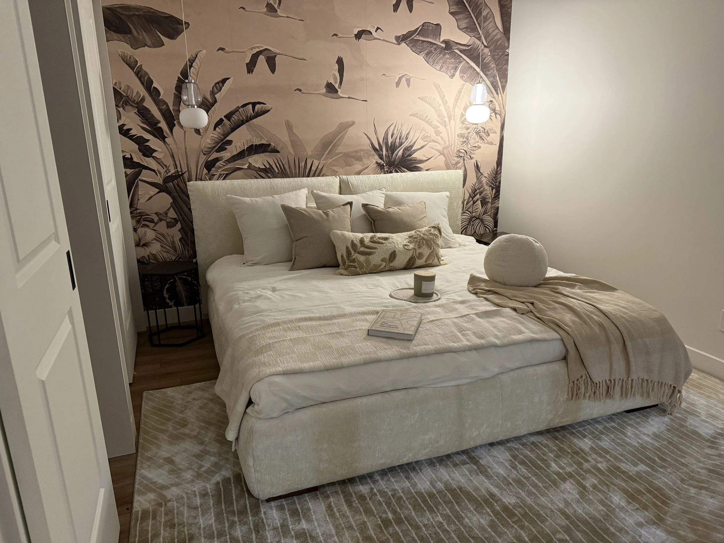 A cozy bedroom with a cream-colored bed and multiple pillows, a textured beige blanket, a decorative round pillow, a small candle, and an open book on the bed. The wall behind the bed features a large tropical leaf wallpaper in muted tones, and there are two modern hanging pendant lights on either side of the bed. The room has a neutral color palette with beige, white, and earthy tones.