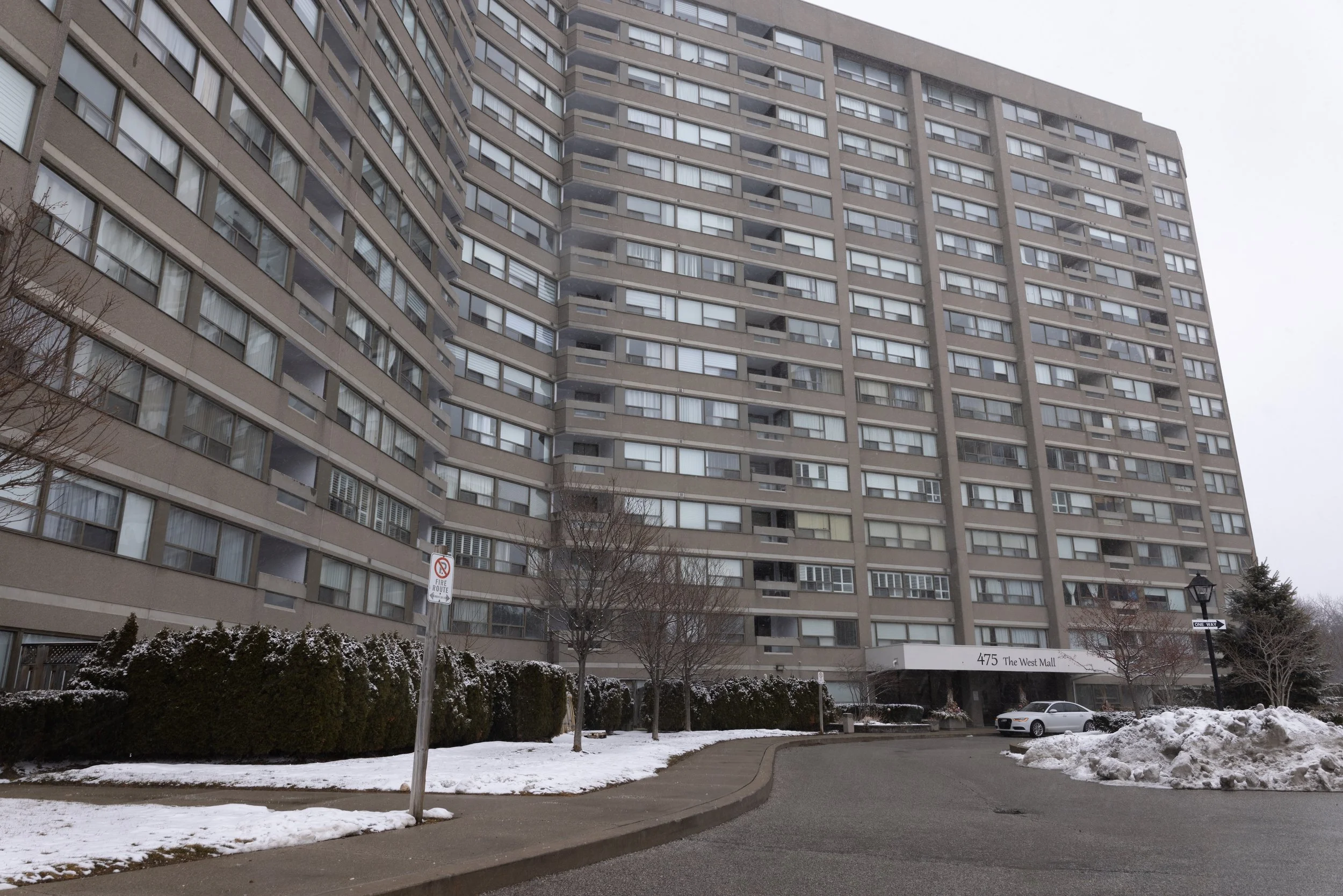 A large residential apartment building with many windows, some balconies, and a gray facade. Snow covers the ground and bushes in the foreground, with a parking lot and a few cars near the building's entrance. There is a sign reading '475 The West Mall' and a streetlamp.