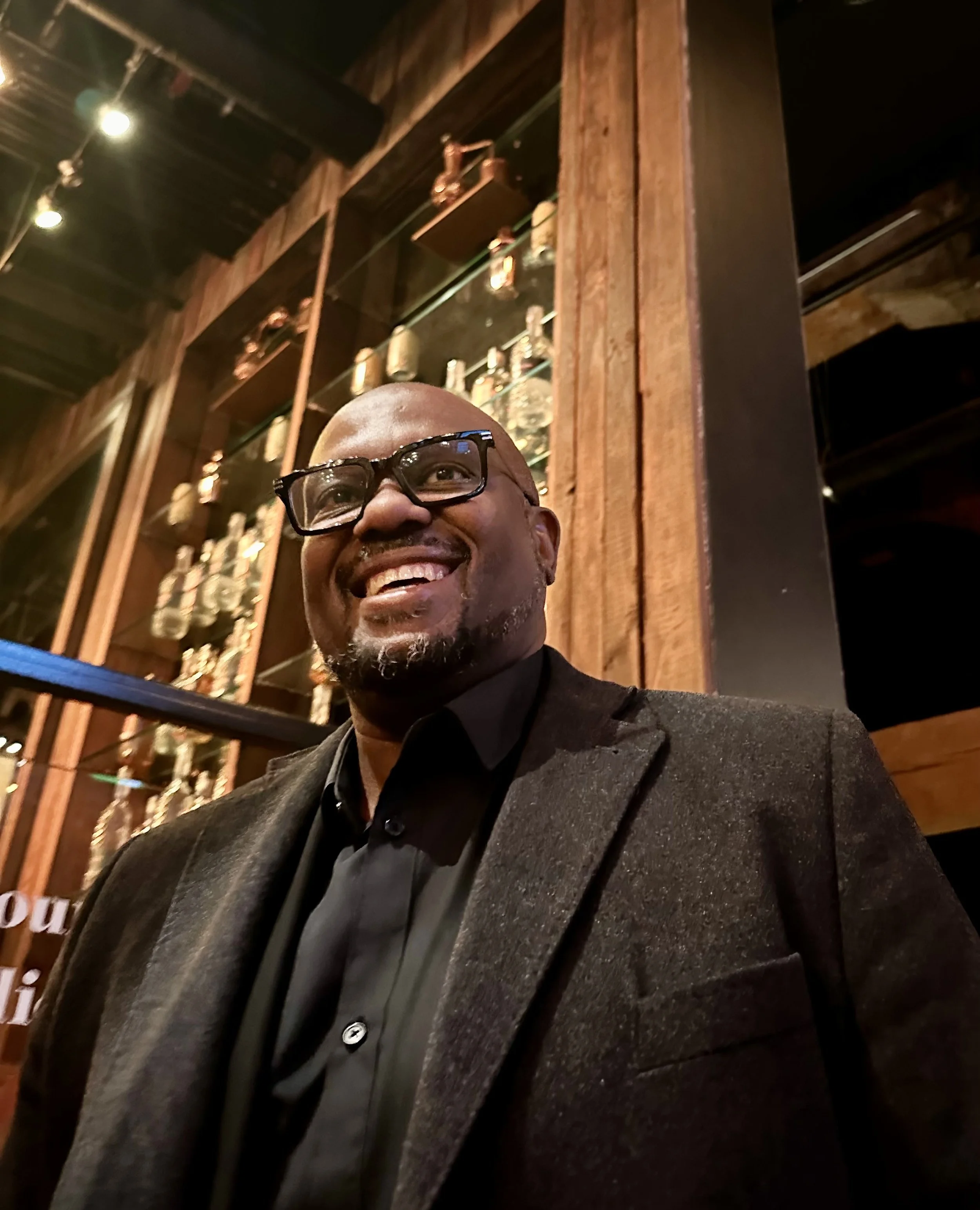 A smiling man with glasses in a dark suit, standing in a dimly lit bar or restaurant with wooden decor and bottles displayed behind him.