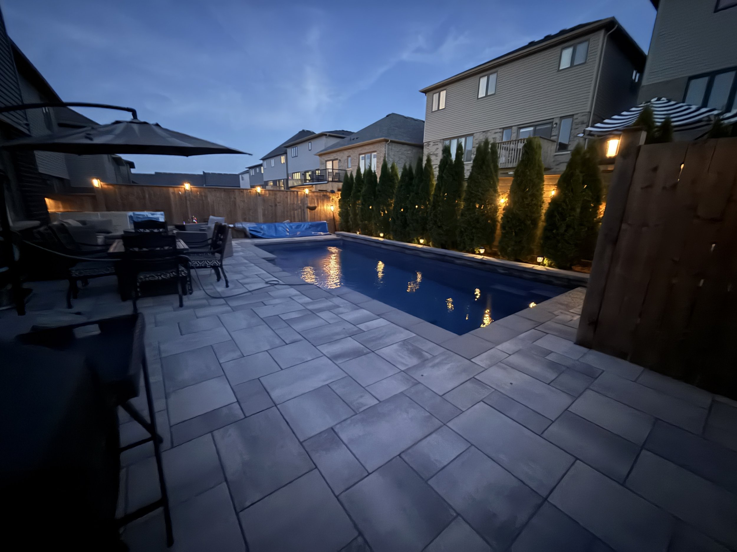 Real Estate Niagara Homes -  Home backyard pool area at dusk with patio furniture, umbrella, heated pool cover, and string lights along the wooden fence, surrounded by neighboring houses.