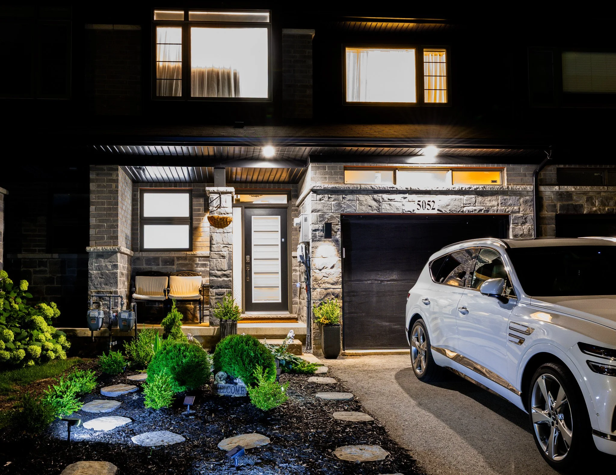 Nighttime exterior view of a modern house with stone and brick facade, illuminated windows, front porch with two chairs, a white SUV parked in the driveway, and a landscaped front yard with lights and plants.