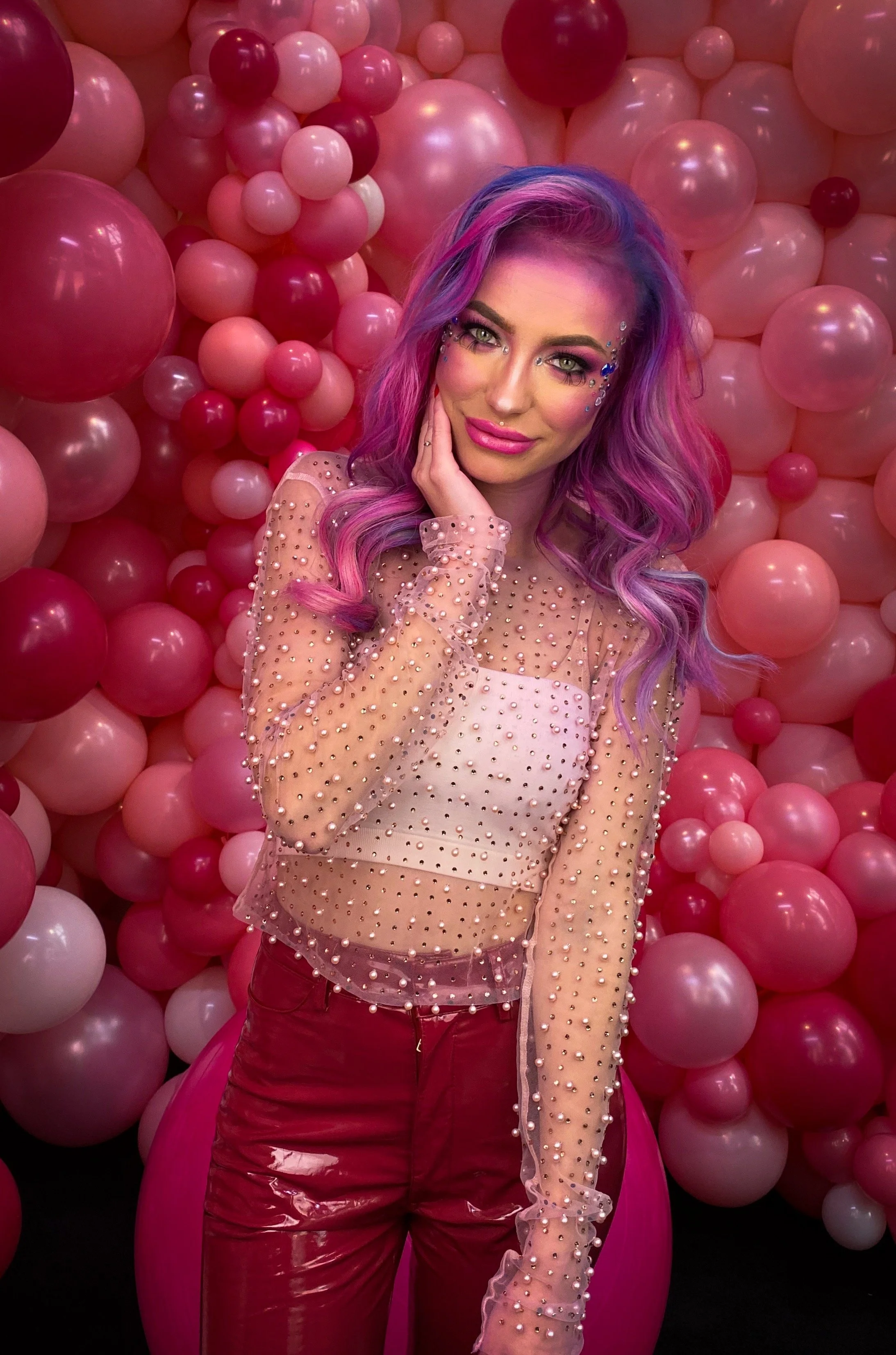 A woman with pink and purple wavy hair posing in front of a pink balloon wall, wearing a sheer pink top with pearl embellishments and red pants.
