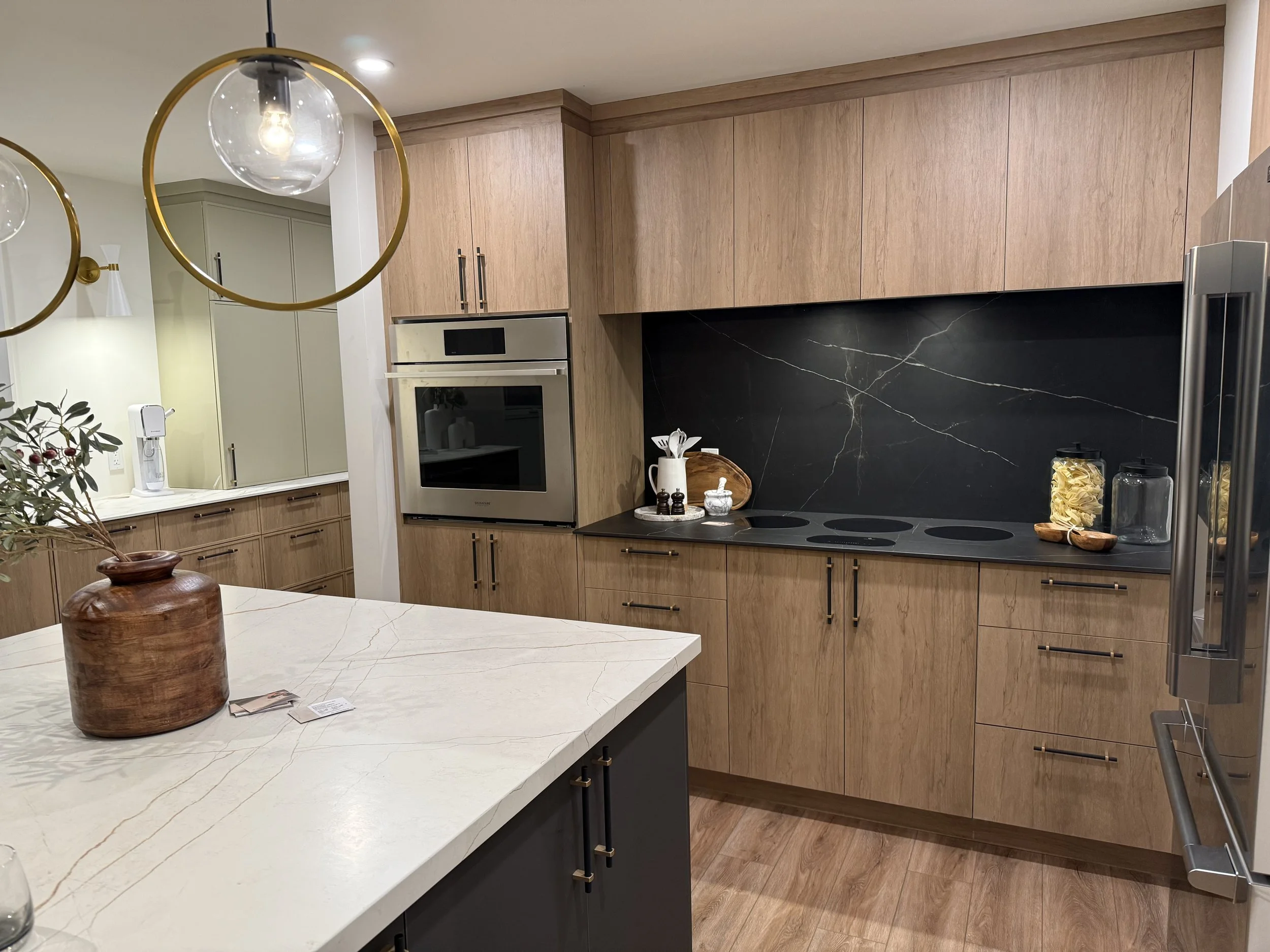 Real Estate Niagara Homes - Modern kitchen with light wood cabinets, black marble backsplash, and a white marble island. There are hanging glass pendant lights, a built-in oven, and various jars and utensils on the countertop.