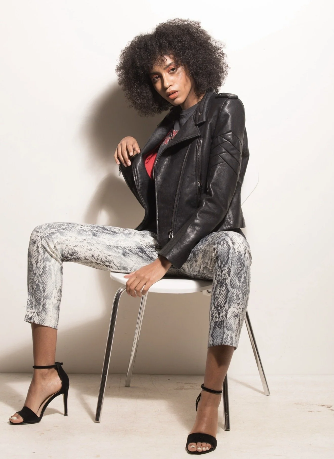 A young woman with curly hair sitting on a white chair against a plain white wall. She wears a black leather jacket, snakeskin print pants, and black high-heeled sandals.