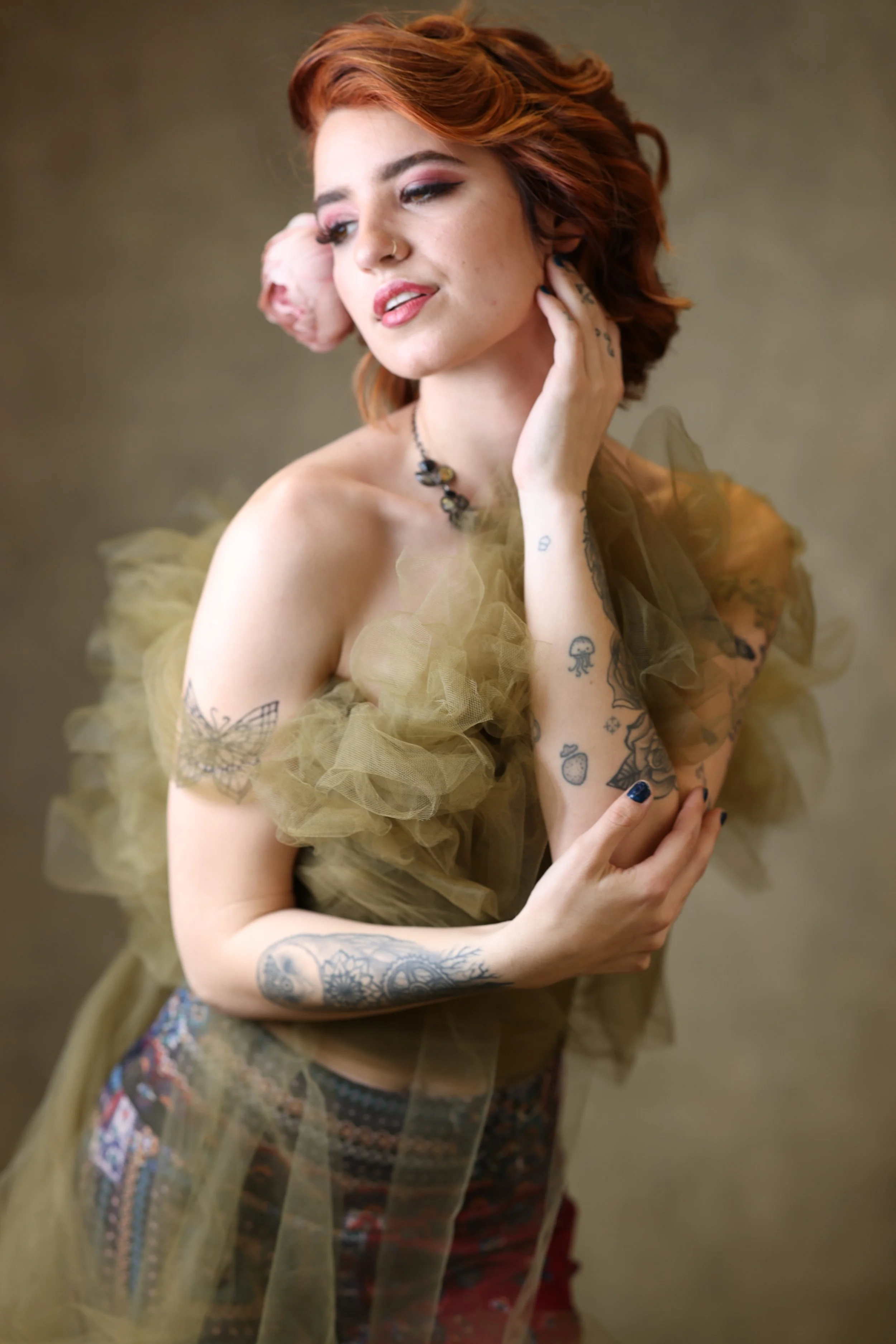A portrait of a woman with red hair, tattoos on her arms, shoulder, and hand, wearing a sheer, ruffled, yellowish dress and a necklace, with a pink flower in her hair. She is touching her face and looking away.