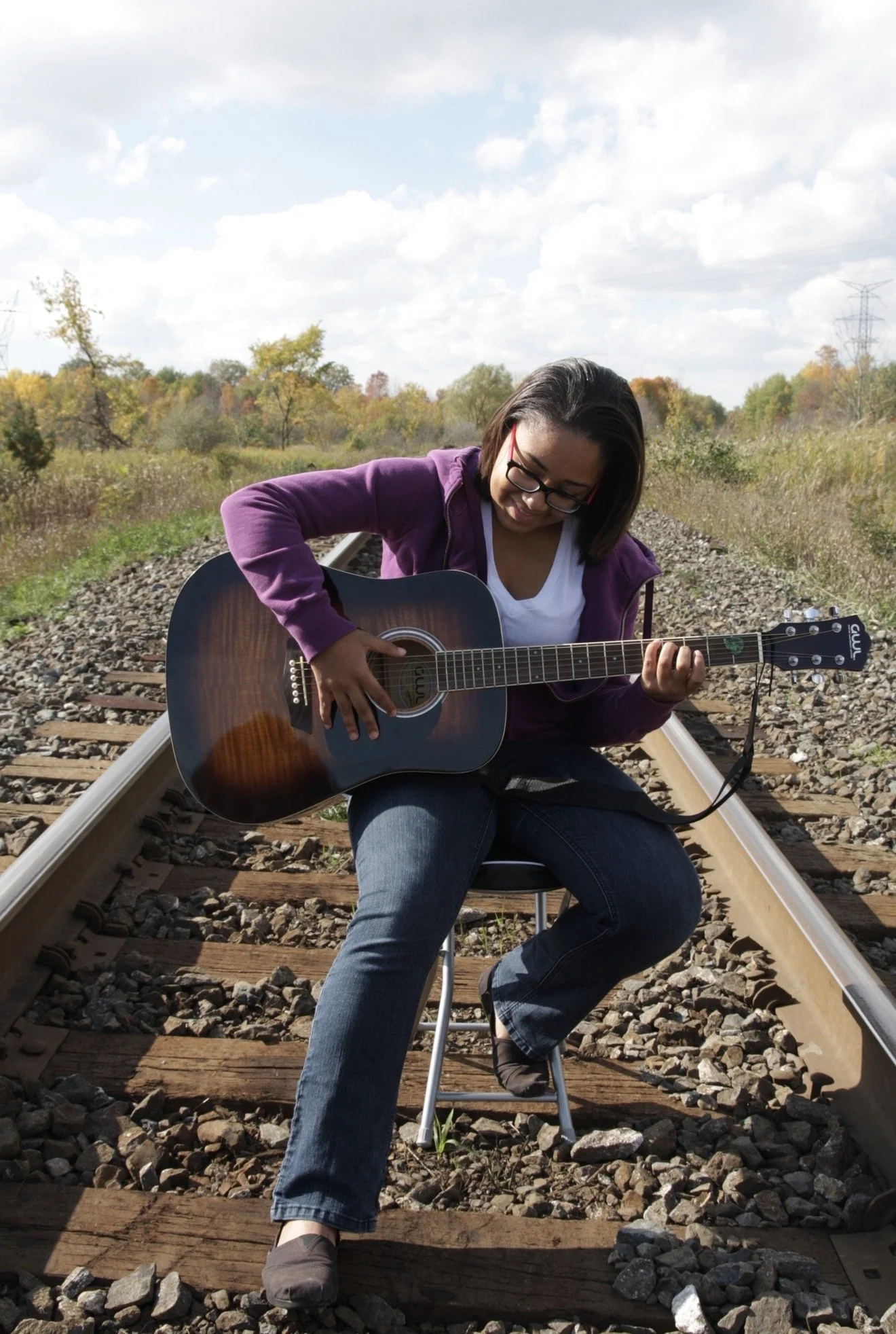 A young woman sitting on a chair playing an acoustic guitar on train tracks outdoors under a partly cloudy sky.