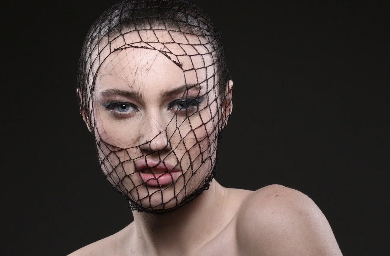 A woman with blue eyes and dark makeup wears a black fishnet covering her face and head, with a black background.