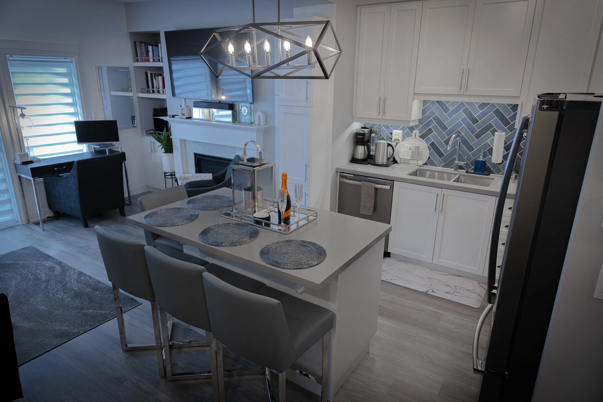 Modern kitchen and living area with white cabinetry, a mosaic tile backsplash, a kitchen island with four barstools, and a dining table with four placemats. There's a fireplace, a small workspace with a computer, and various appliances including a coffee maker and refrigerator.