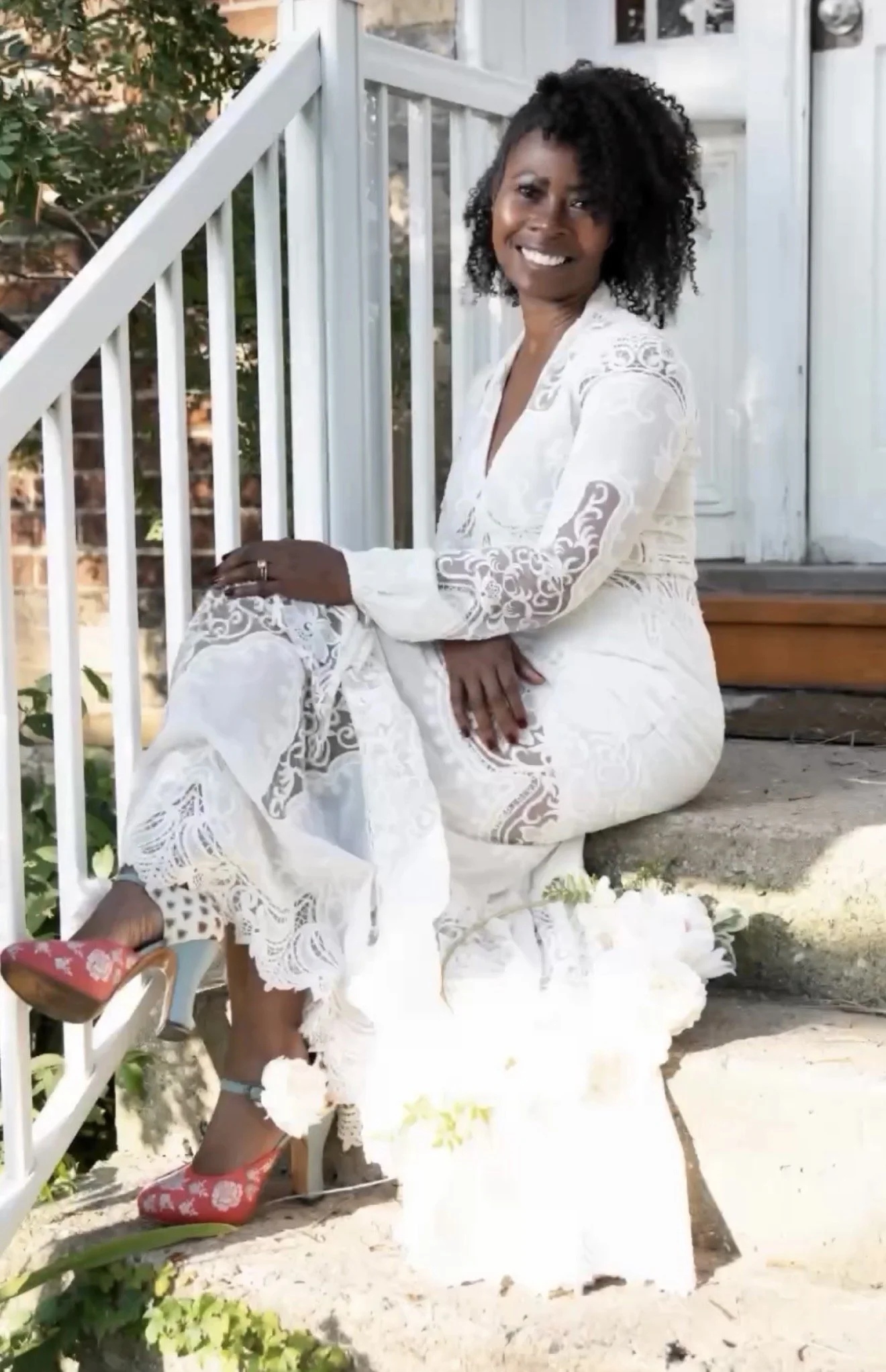 A woman dressed in a white lace dress with a floral pattern, sitting on steps outside a house, smiling and looking at the camera, wearing colorful high heels, with a bouquet of white flowers nearby.