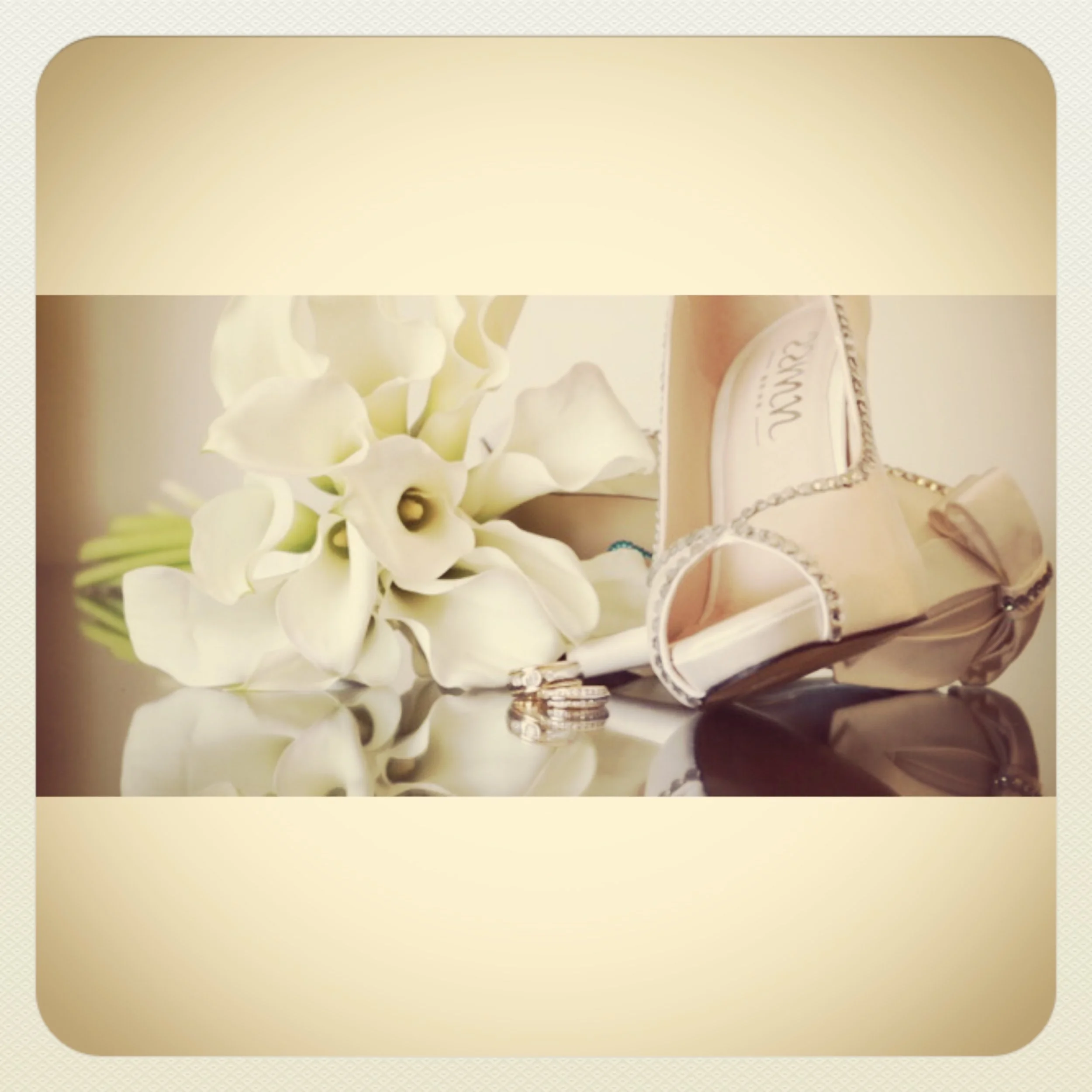 A wedding bouquet of white calla lilies, wedding rings, a white high-heeled shoe with rhinestone embellishments, and a small purse, all displayed on a reflective surface.