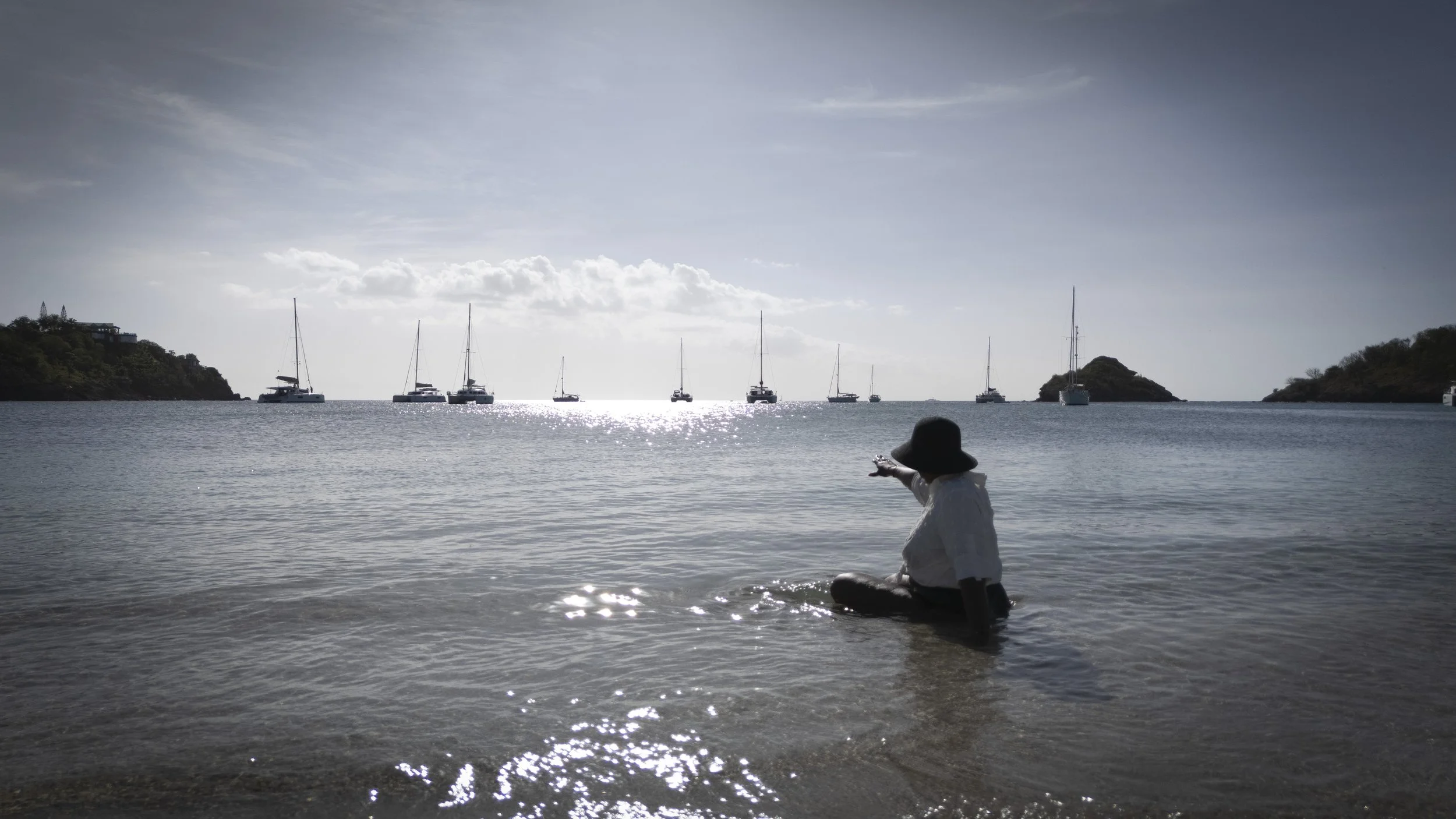 A person sitting in shallow water on a beach, pointing towards sailboats anchored in the bay with an island and cloudy sky in the background.