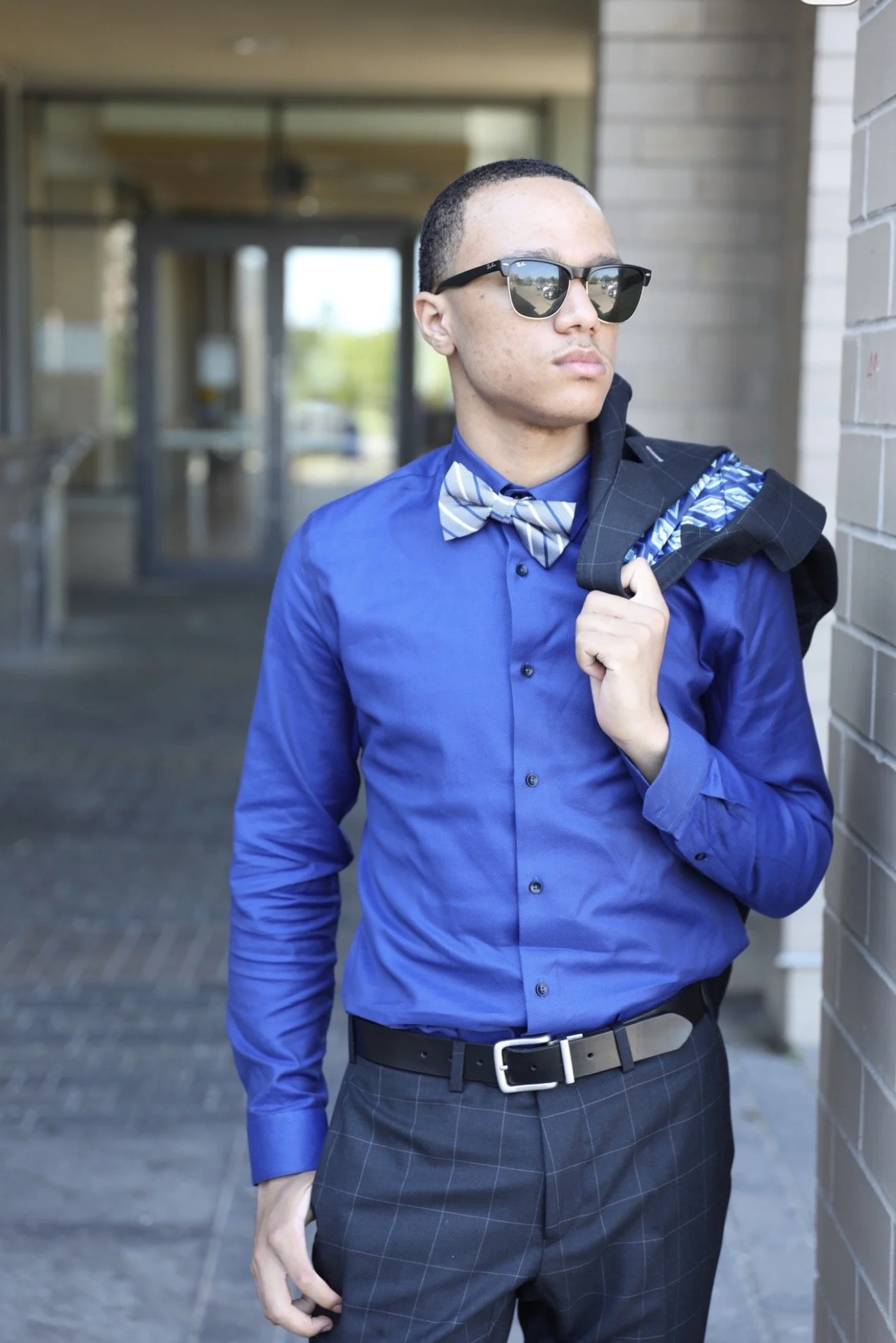 Young man wearing a blue dress shirt, plaid bow tie, and sunglasses, holding a jacket over his shoulder, standing outside against a brick wall.