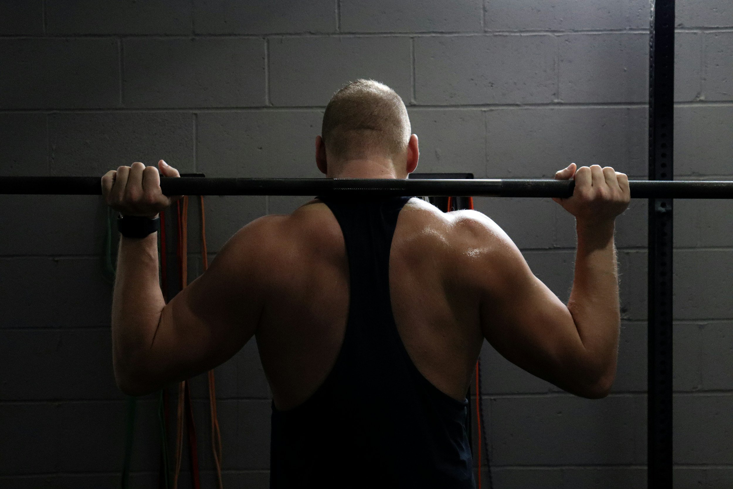 A man with muscular arms and a shaved head, wearing a black sleeveless tank top, is lifting a barbell at the gym. He is facing a gray brick wall with exercise bands hanging on the side.