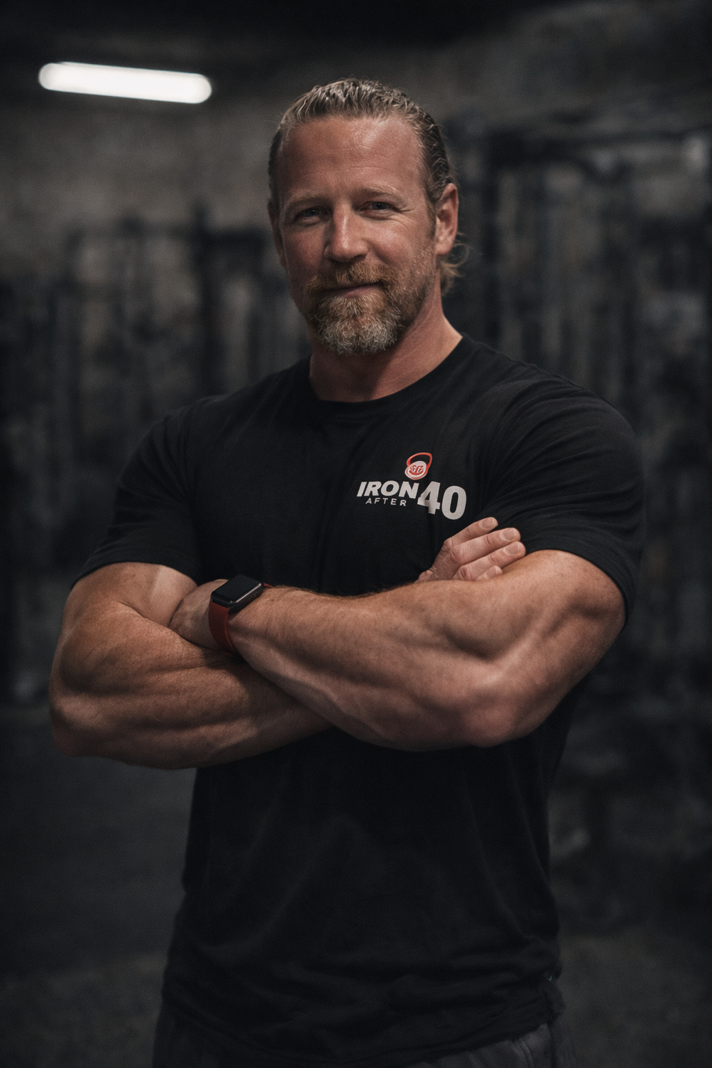 A confident middle-aged man with a beard and long hair tied back, standing with arms crossed in a gym, wearing a black T-shirt with the words 'Iron 40' and a smartwatch on his wrist.