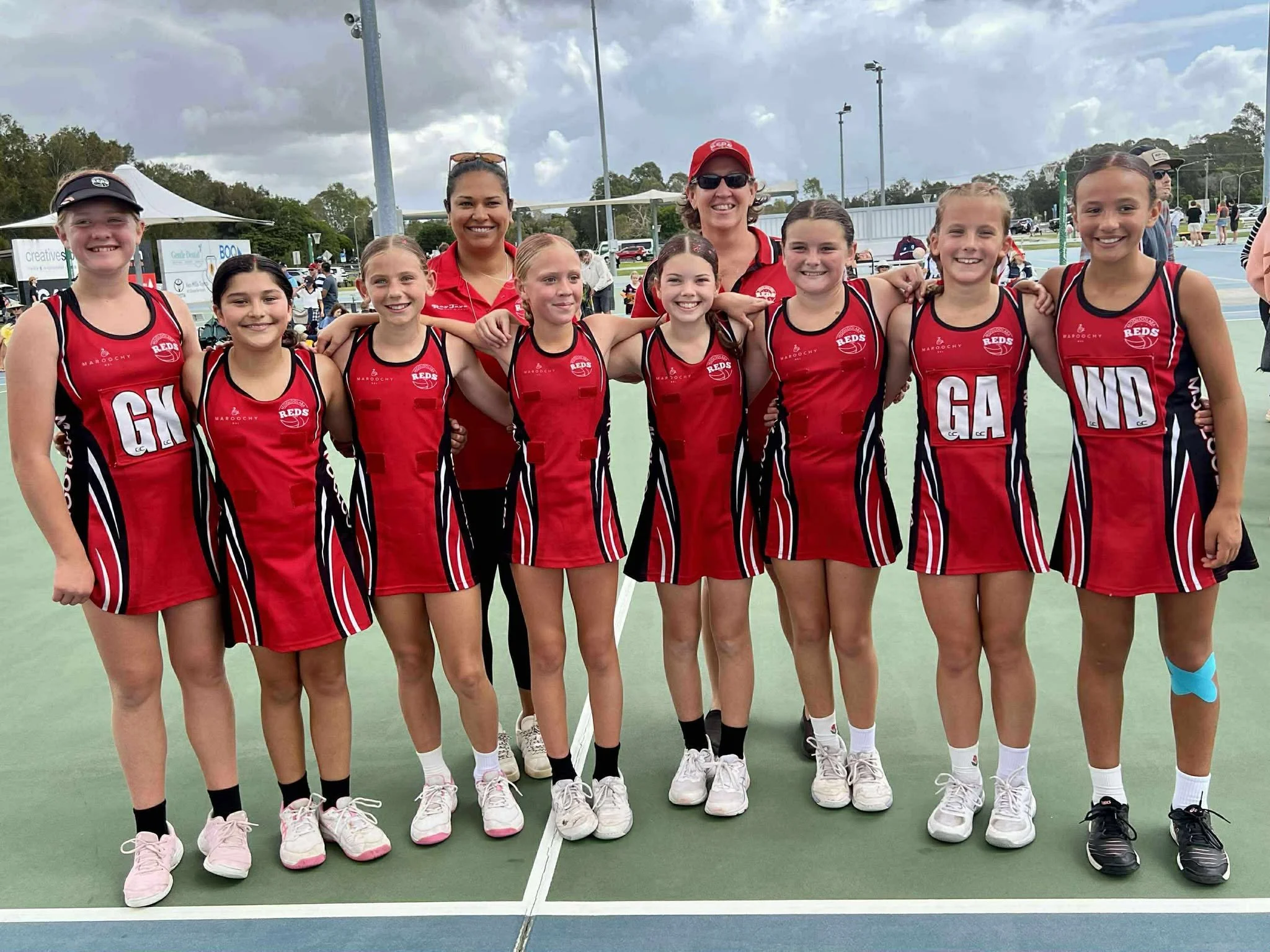 Mooloolaba Reds team and their coaches standing on a netball court, at Fishermans Road Netball