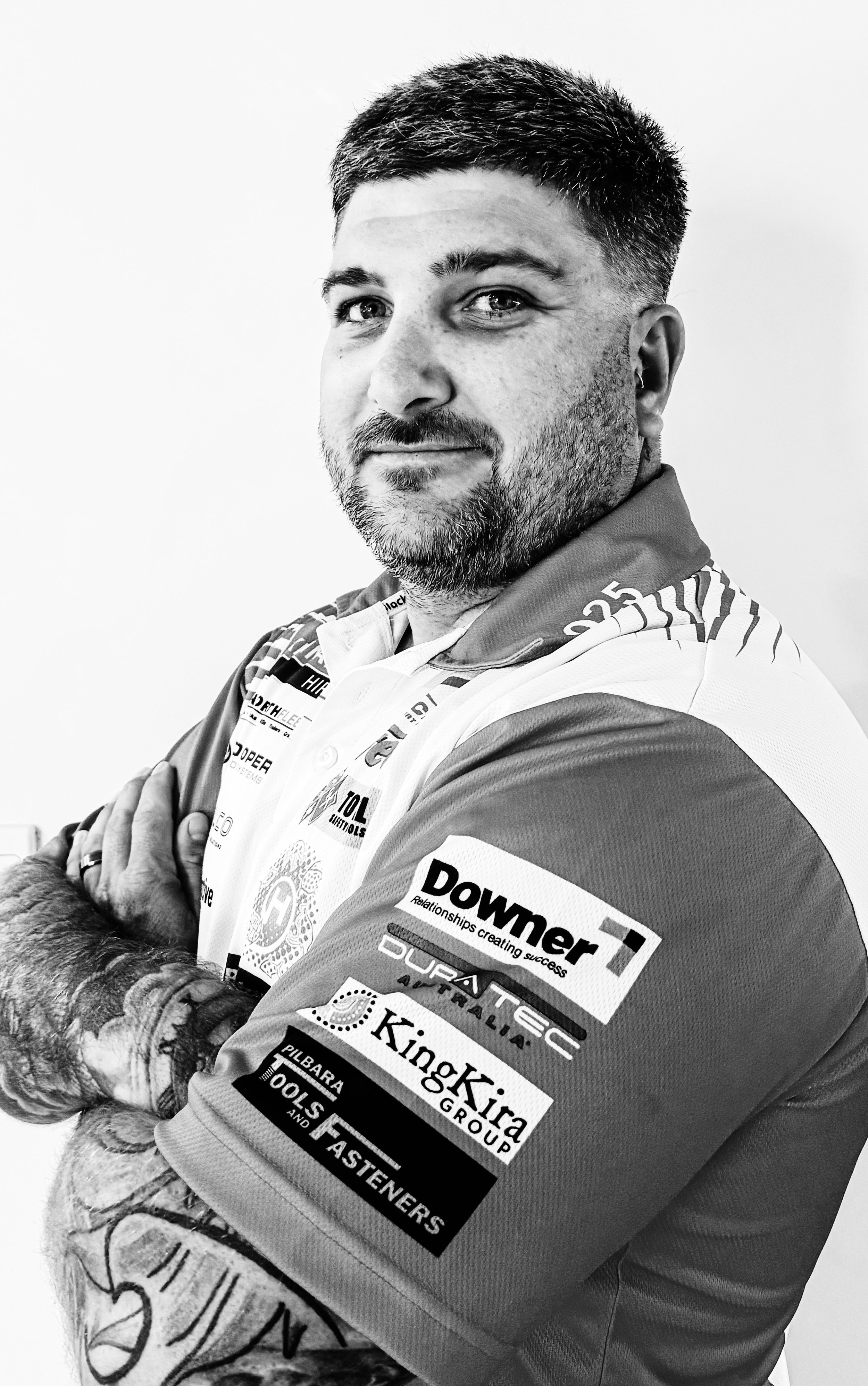 Black and white photo of a man with short hair and a beard, wearing a racing jersey with various sponsor logos, crossing his arms and looking at the camera.