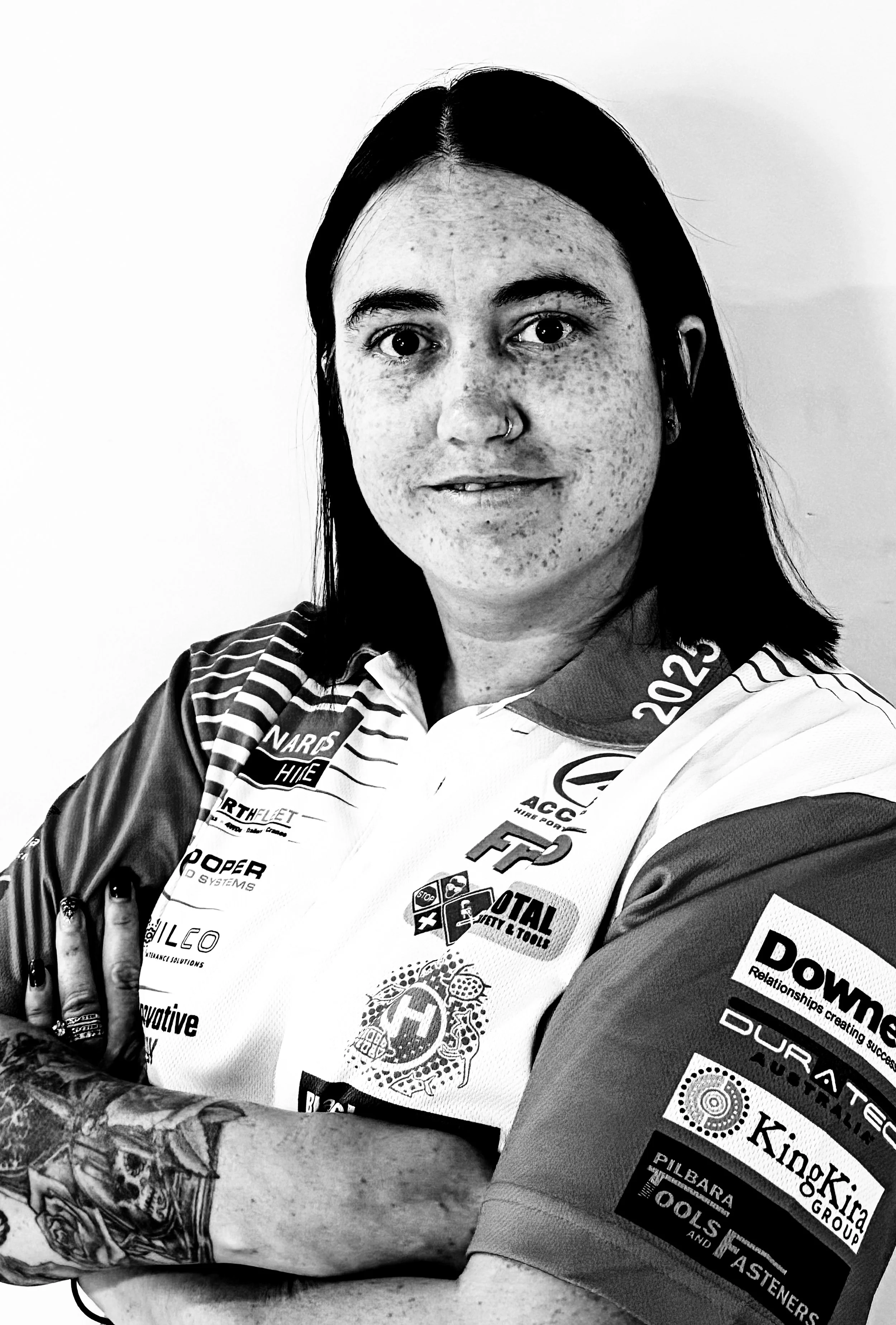 A woman with dark hair, tattoos, and a nose ring wearing a racing team jersey with various sponsor logos, smiling with her arms crossed.